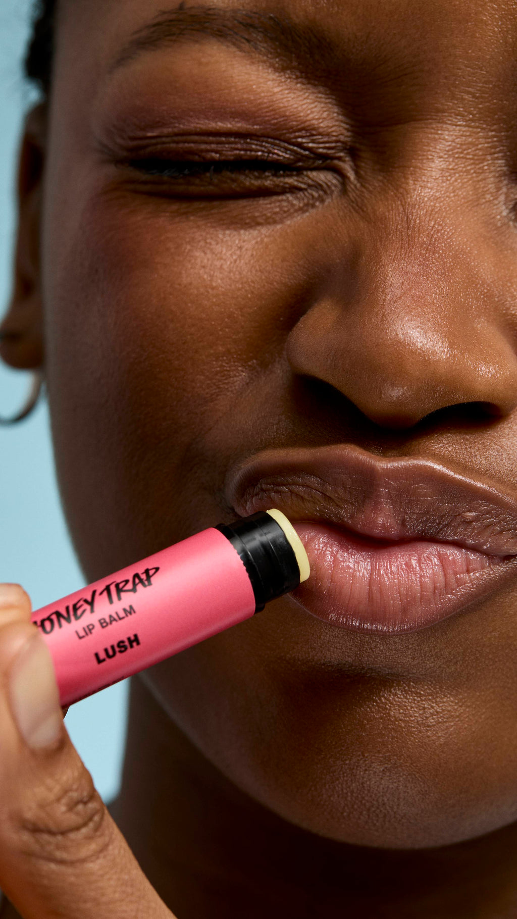 A close-up of a person applying pink 'Honey Trap' lip balm to their lips. They have their eyes closed with a playfully scrunched expression, highlighting the balm's application against a soft blue background.
