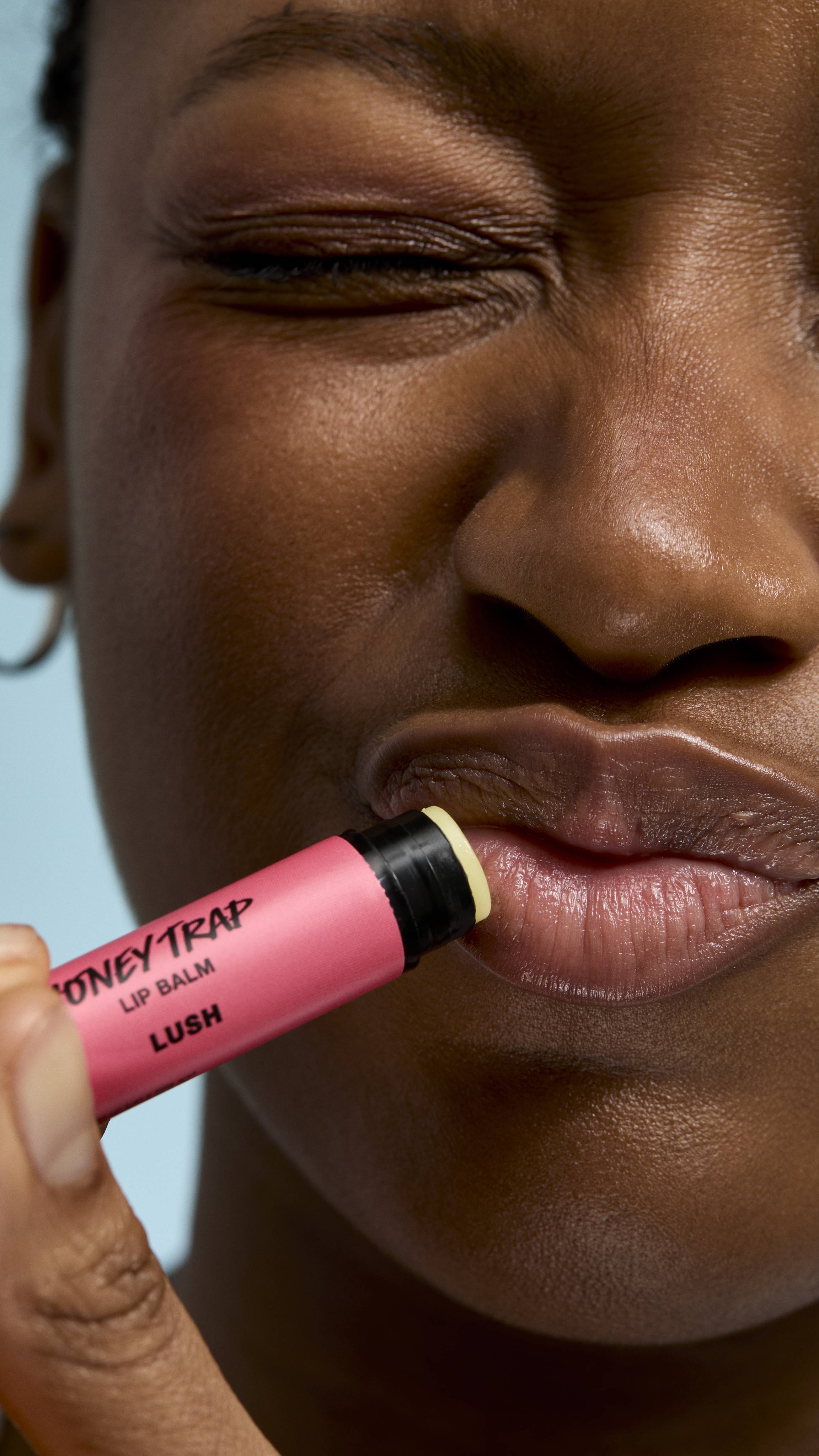 A close-up of a person applying pink 'Honey Trap' lip balm to their lips. They have their eyes closed with a playfully scrunched expression, highlighting the balm's application against a soft blue background.