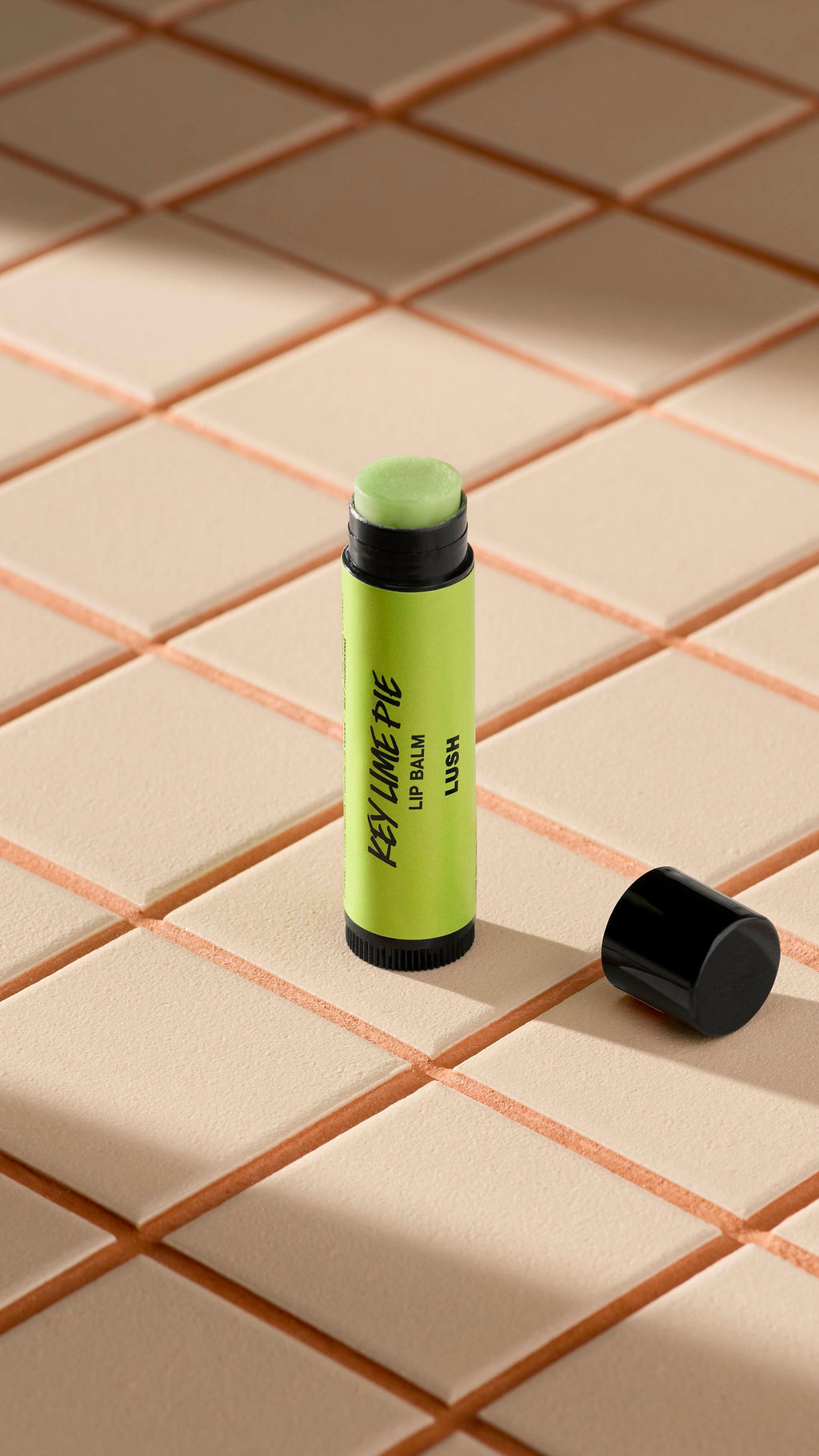 A green-labelled tube of Lush 'Key Lime Pie' lip balm stands upright on light-coloured square tiles. The black cap is off, lying next to the base, revealing the pale, creamy balm inside. Soft shadows fall across the tiled surface.