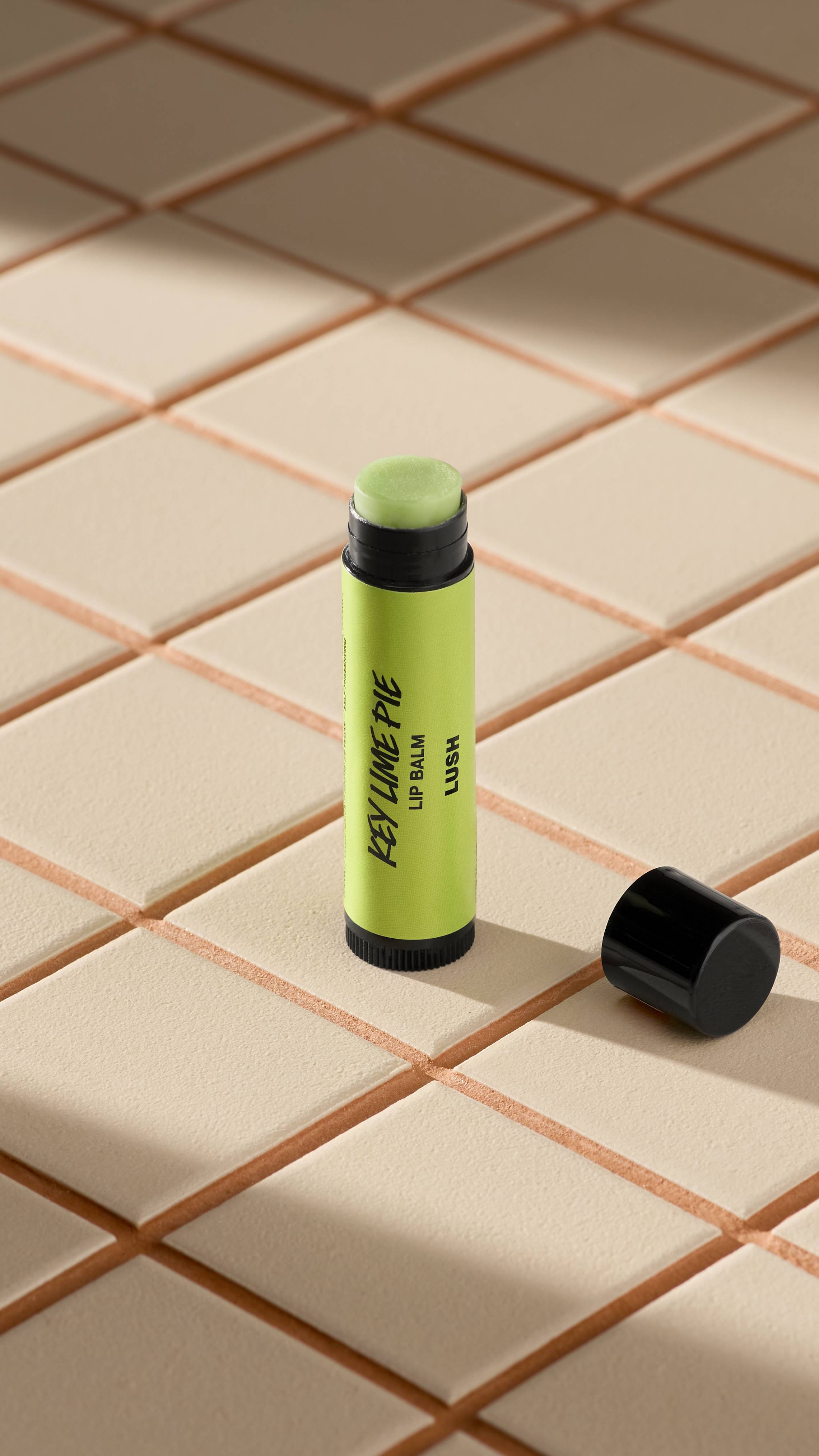 A green-labelled tube of Lush 'Key Lime Pie' lip balm stands upright on light-coloured square tiles. The black cap is off, lying next to the base, revealing the pale, creamy balm inside. Soft shadows fall across the tiled surface.
