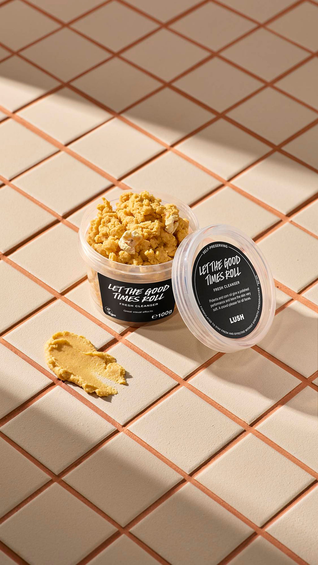 Open tub of Lush 'Let the Good Times Roll' cleanser and a yellow swatch on light beige tiles with brown grout. The tub is full of the crumbly product and features black product labels.