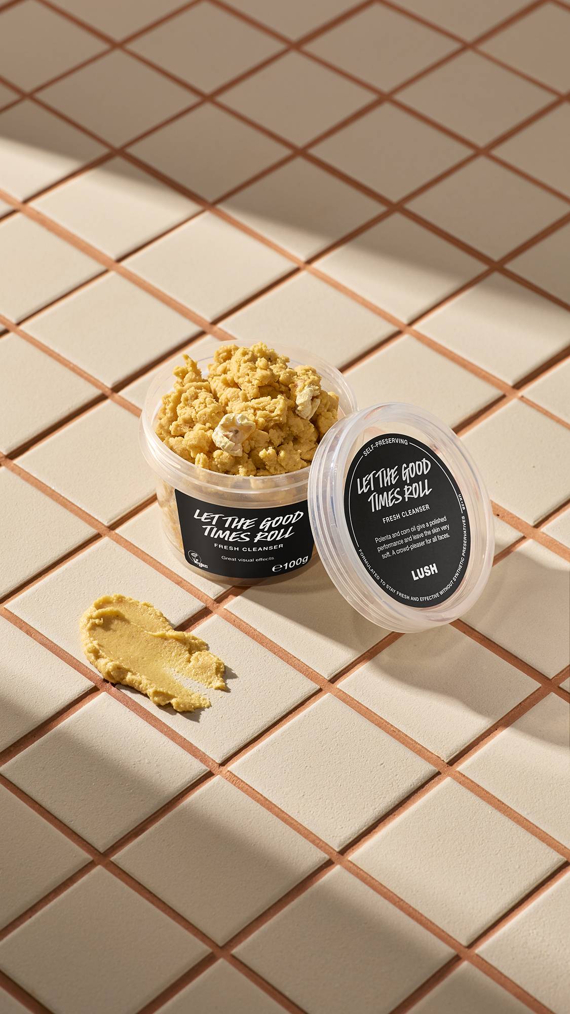 Open tub of Lush 'Let the Good Times Roll' cleanser and a yellow swatch on light beige tiles with brown grout. The tub is full of the crumbly product and features black product labels.