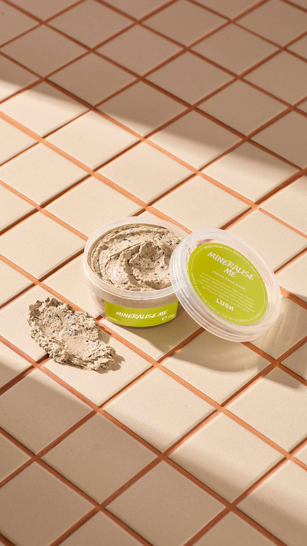 The image shows a clear, recycled plastic, open pot of Mineralise Me face mask with lime-green labels, the lid leaning on the side and a smeared swatch of the textured product on the cream-tiled floor beside the pot.