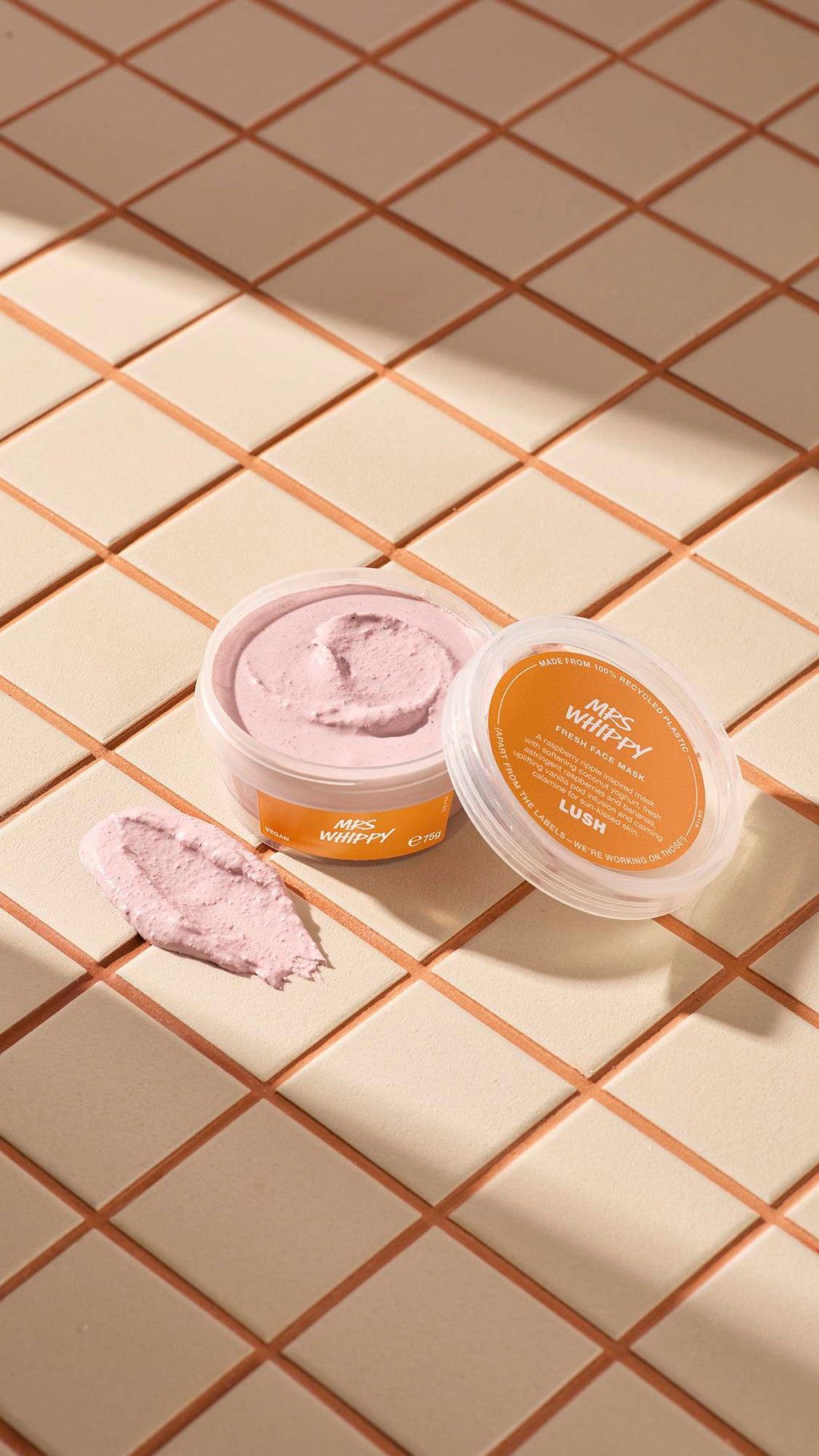 The image shows a clear, recycled plastic, open pot of Mrs Whippy face mask with orange labels, the lid leaning on the side and a smeared swatch of the textured product on the cream-tiled floor beside the pot.