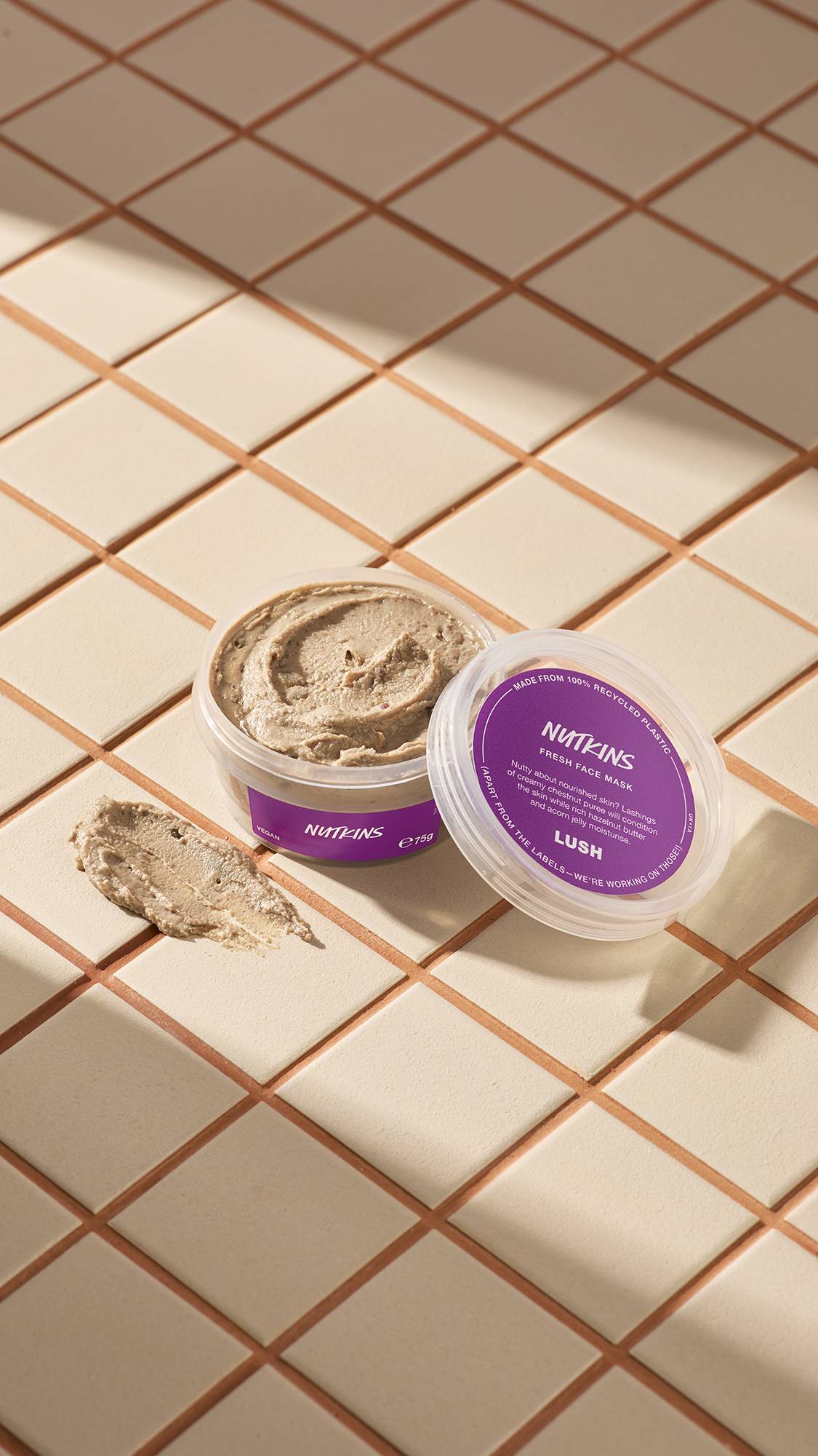 The image shows a clear, recycled plastic, open pot of Nutkins face mask with purple labels, the lid leaning on the side and a smeared swatch of the textured product on the cream-tiled floor beside the pot.