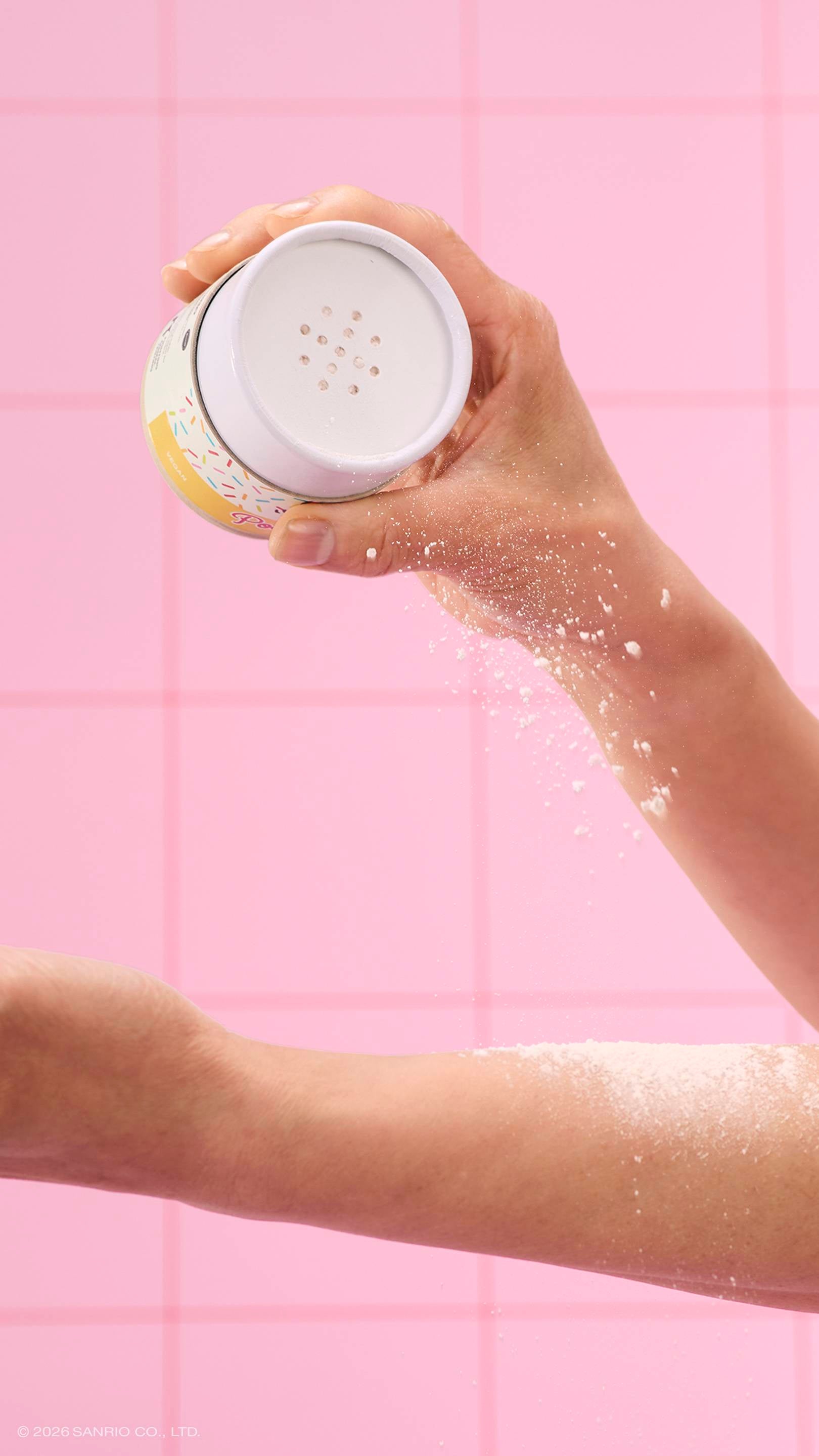 A close-up of a model's hand shaking white dusting powder from a Lush x Pochacco tube onto their other arm. The scene is set in a pink tiled bathroom.