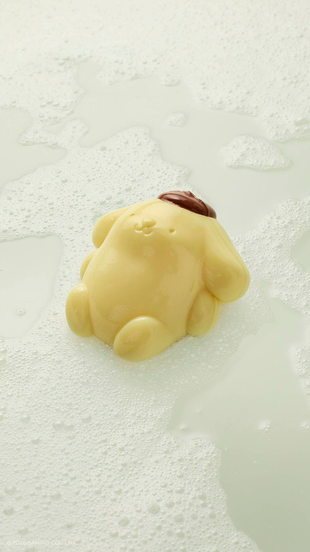 A glossy, pale yellow Pompompurin shower jelly rests in a pool of white soapy suds and bubbles. The character, wearing its signature brown beret, is shown from a high-angle view on a light, neutral surface.
