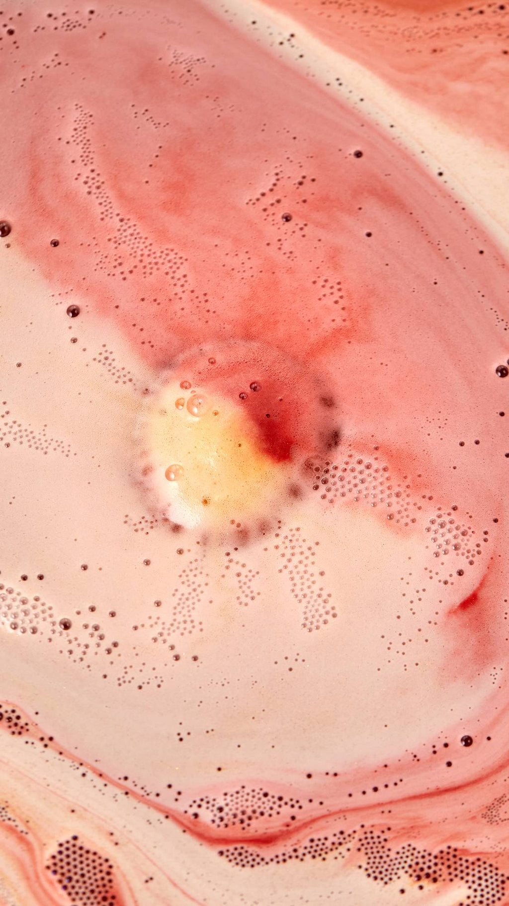 The image shows an aerial view of the Sticky Dates bath bomb as it dissolves in the bath. The product creates a thick blanket of warm, rich, rust-coloured, velvety foam and contrasting ivory foam alongside flecks of glitter.