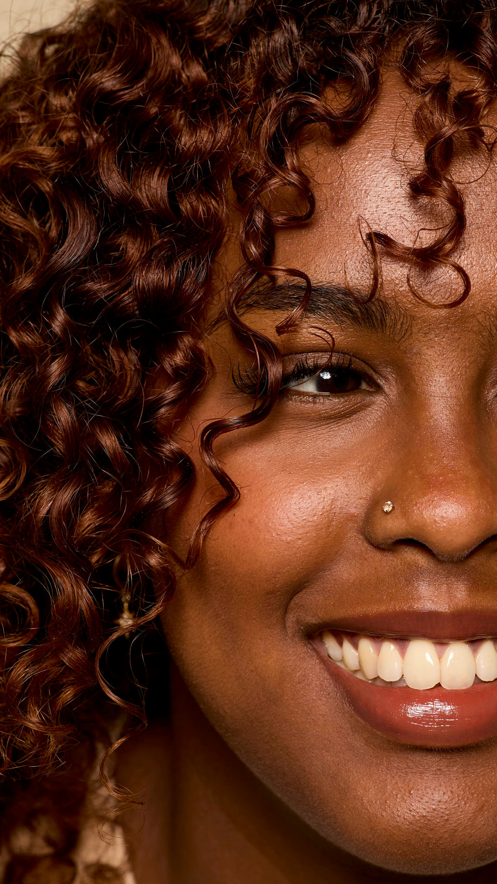 A close-up of a smiling person with defined, light brown curls framing their face. Their skin is dewy and glowing, and a single piercing is visible on their nose, capturing a radiant and joyful moment.