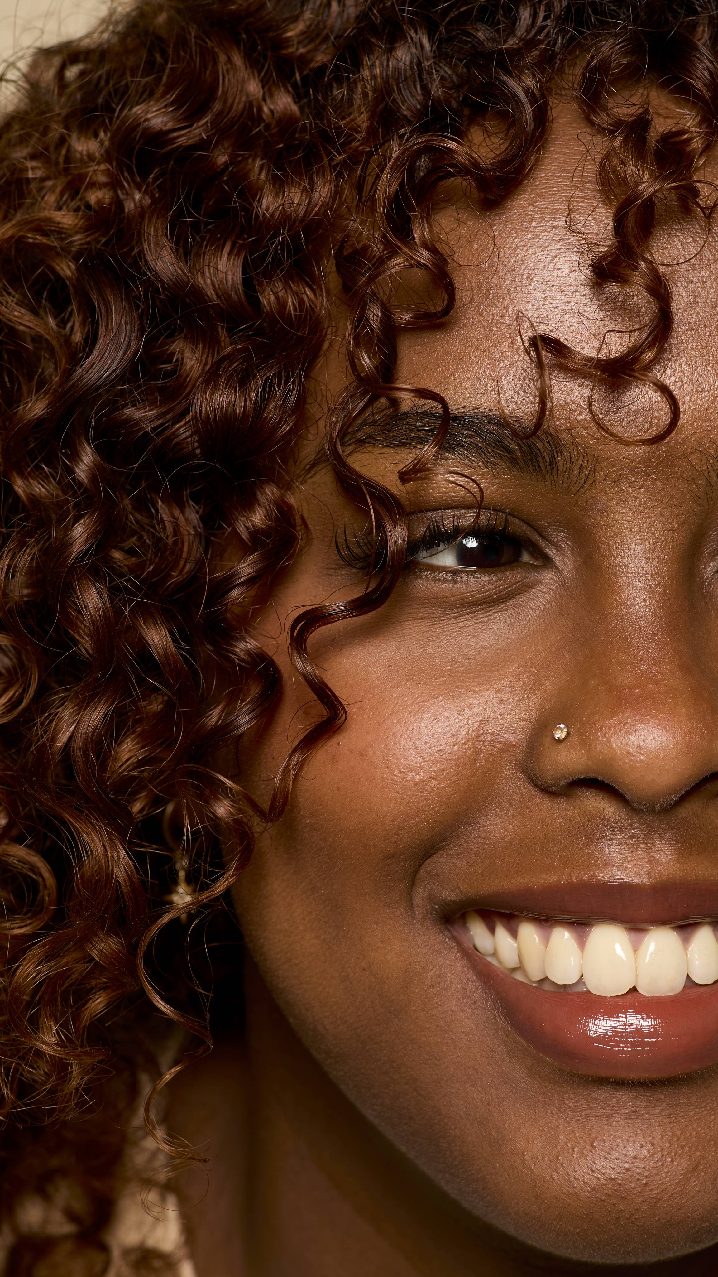 A close-up of a smiling person with defined, light brown curls framing their face. Their skin is dewy and glowing, and a single piercing is visible on their nose, capturing a radiant and joyful moment.