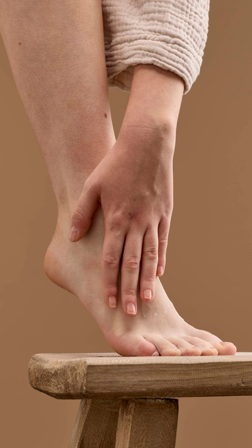 The image shows a hand rubbing a pale cream 'T For Toes' foot powder onto a bare foot and ankle. The foot rests on a wooden stool, with the person's robe visible against a solid beige background.