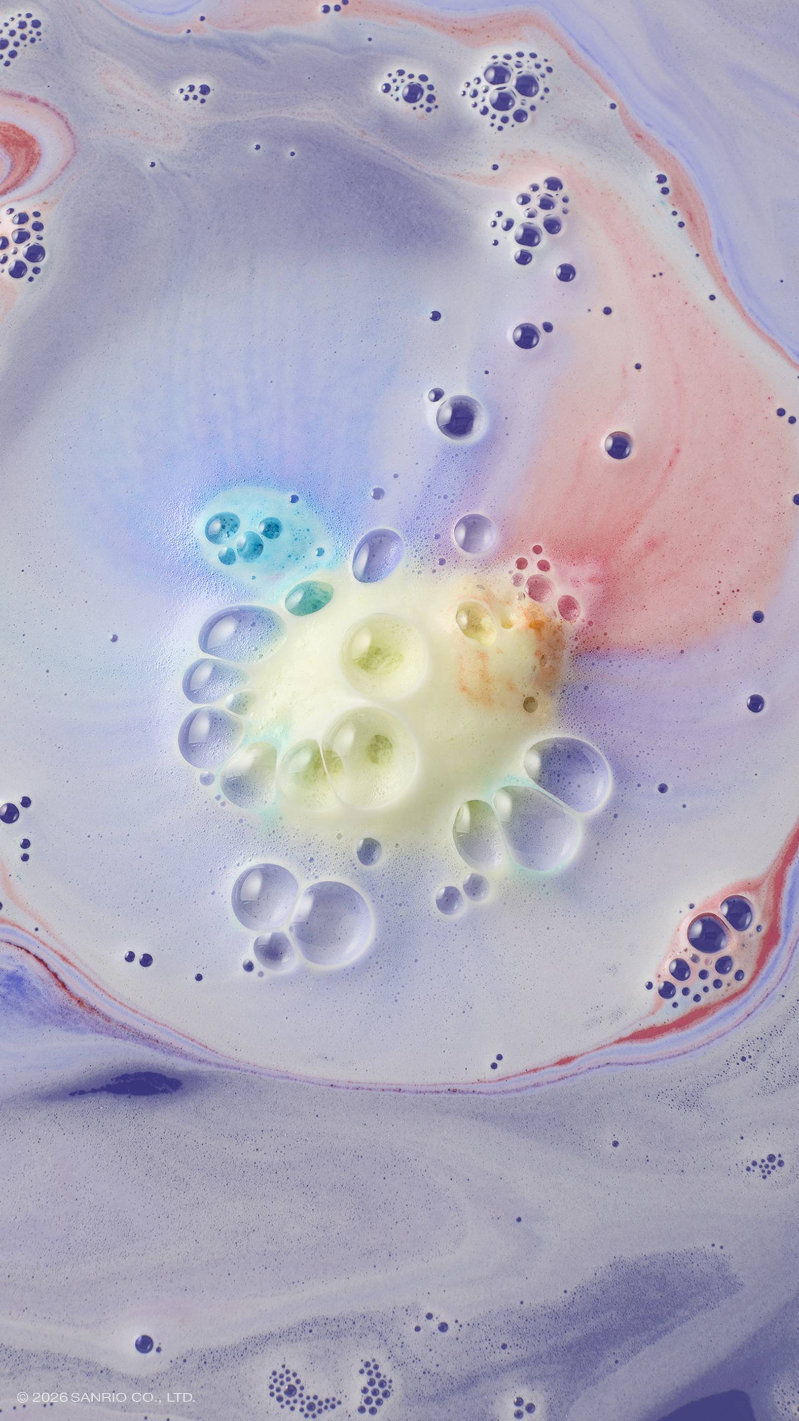 An aerial shot of lavender bath water swirling with multi-coloured foam. In the centre, a cluster of yellow and blue bubbles marks where the "U.SA.HA.NA" bath bomb has dissolved, with soft streaks of pink and purple radiating across the surface.