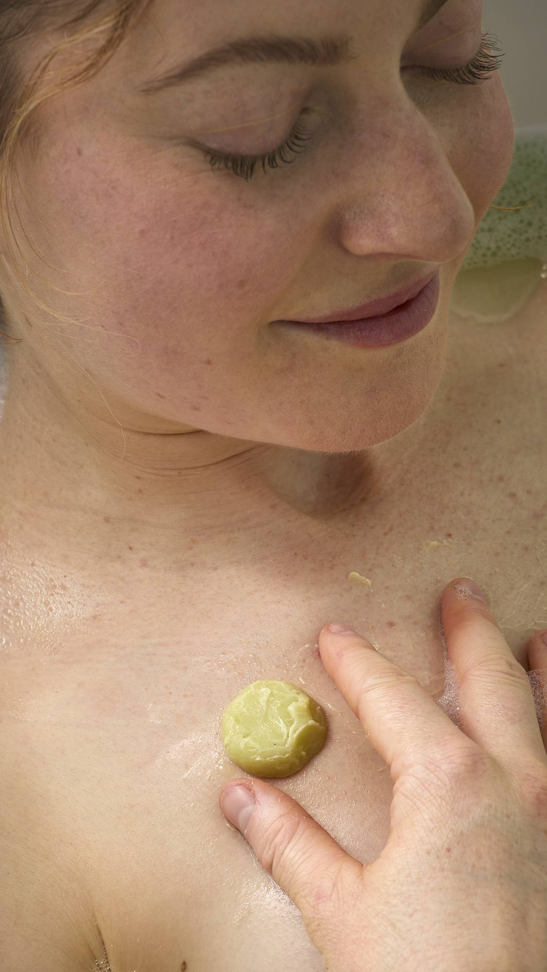 A smiling person relaxes in the bath, massaging the small, moisturizing melt from the Cold Bath Bomb onto their chest. The yellowish-green butter glides across their wet skin, offering a moment of soothing hydration.