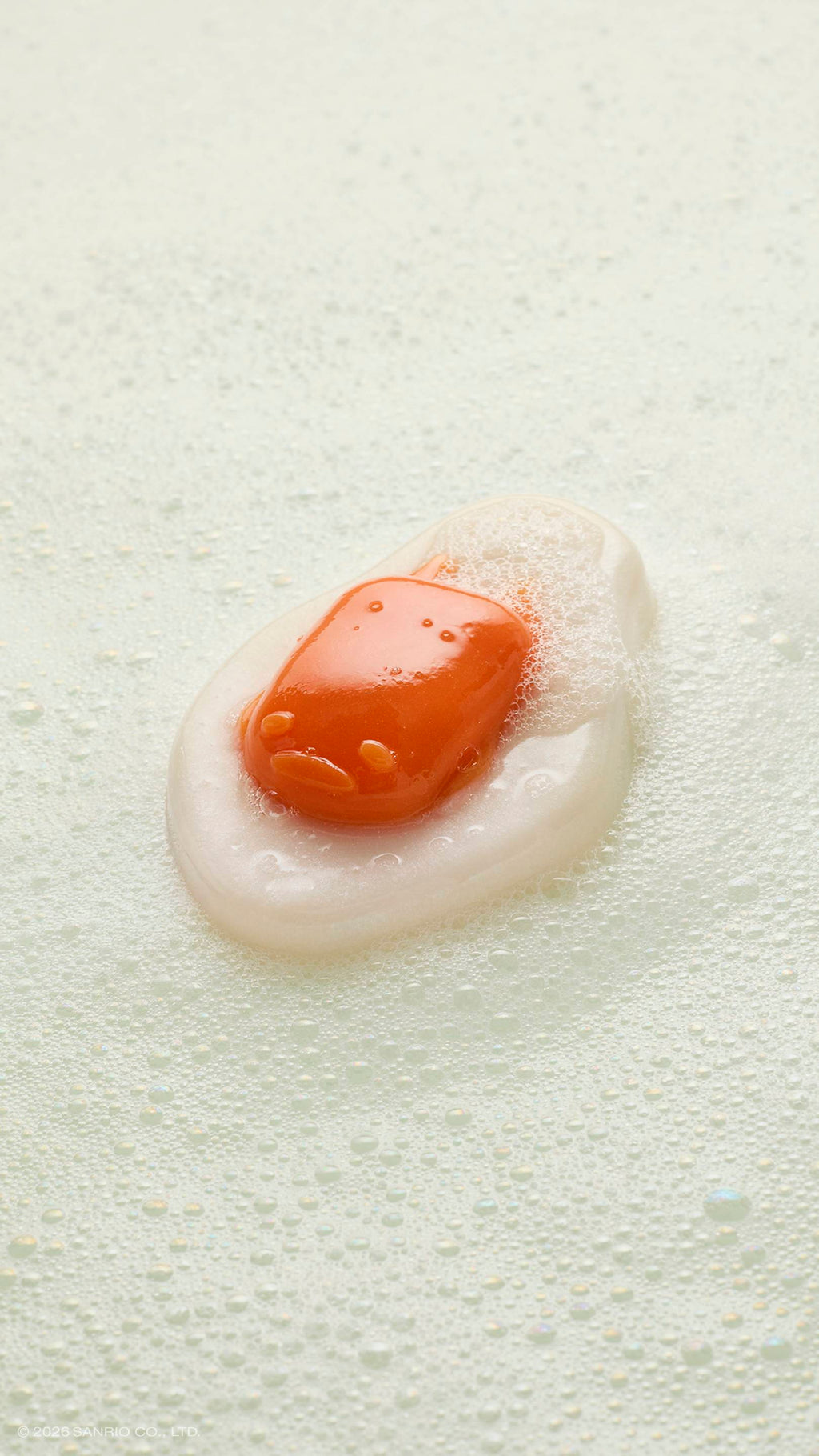 A glossy, fried-egg-shaped shower gummy rests in a pool of white soapy suds and bubbles. The orange "yolk" in the centre features the faint, sleepy face of the character Gudetama.
