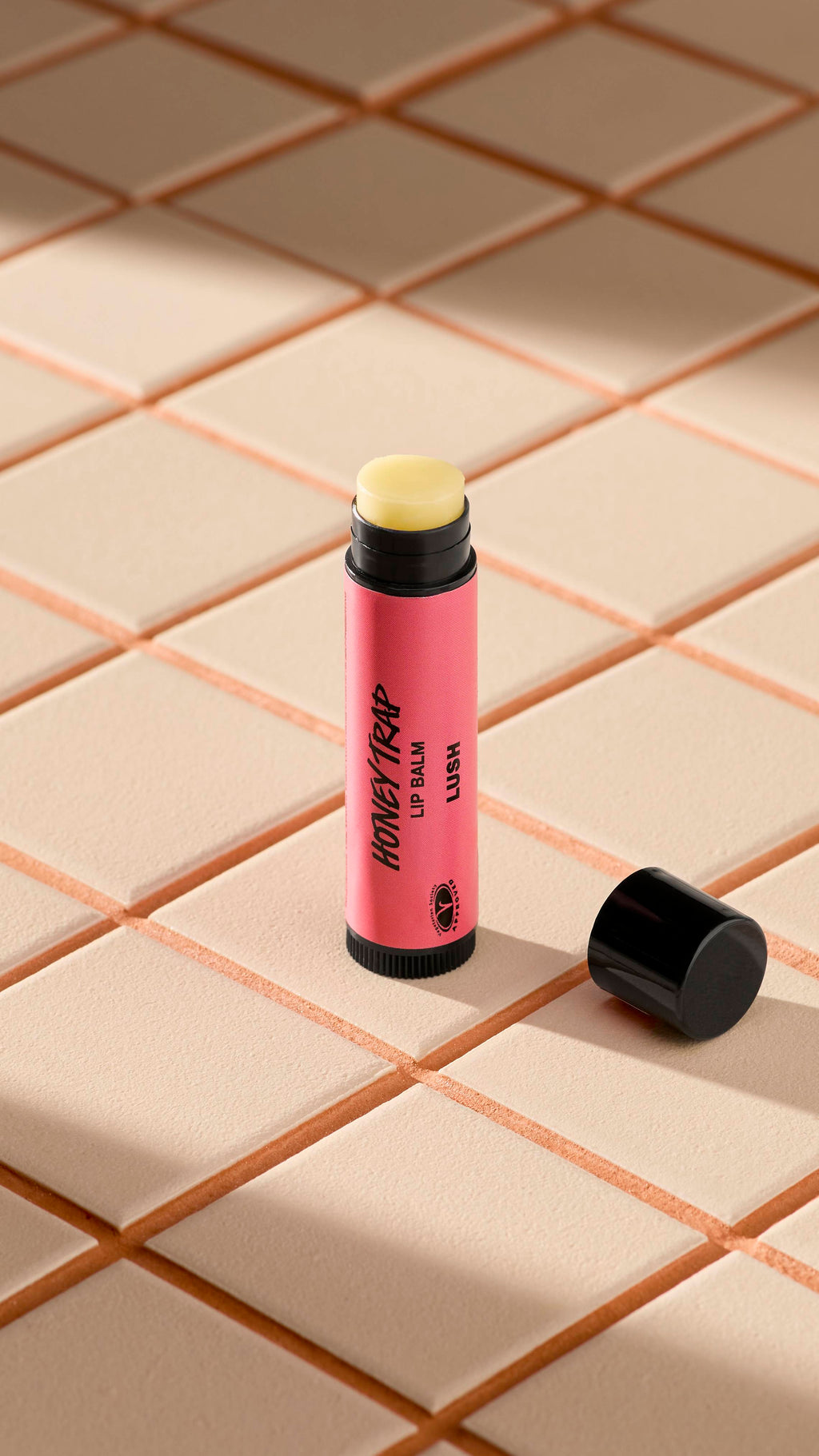 A pink tube of Lush 'Honey Trap' lip balm stands upright on light-coloured square tiles. The black cap is off, lying next to the base, revealing the pale, creamy balm inside. Soft shadows fall across the tiled surface.