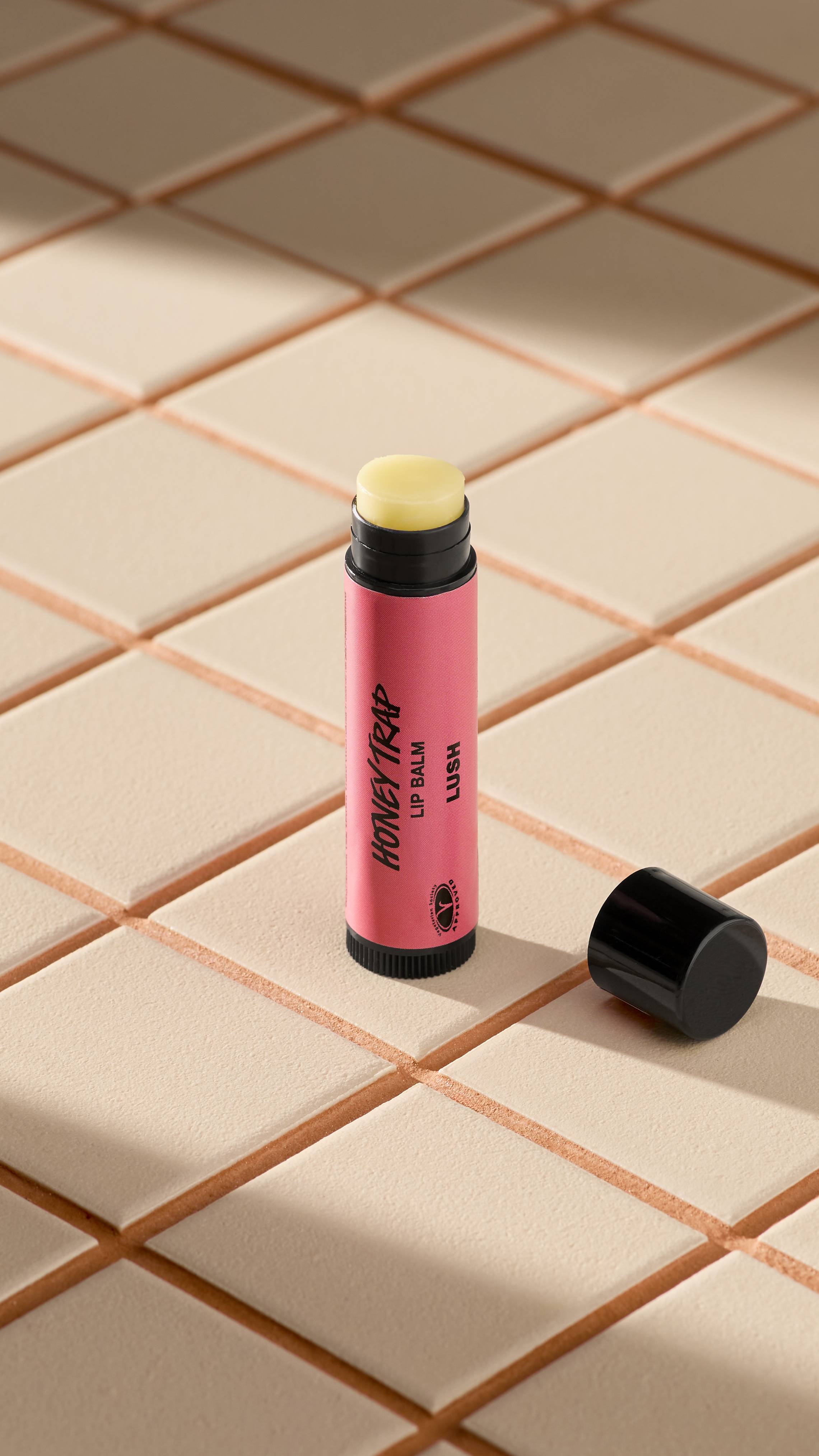 A pink tube of Lush 'Honey Trap' lip balm stands upright on light-coloured square tiles. The black cap is off, lying next to the base, revealing the pale, creamy balm inside. Soft shadows fall across the tiled surface.