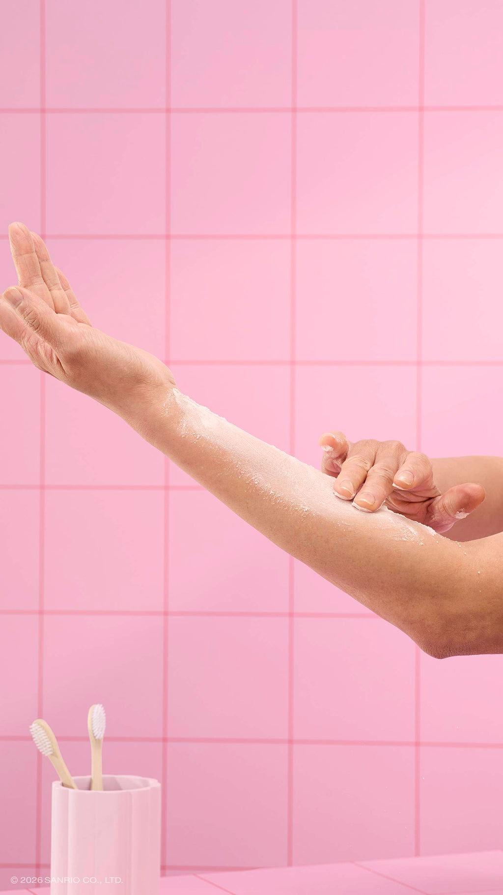 A close-up shows hands gently rubbing white dusting powder into an arm. The scene is set against a background of pink square tiles, with a pink toothbrush holder containing two toothbrushes in the lower-left corner.