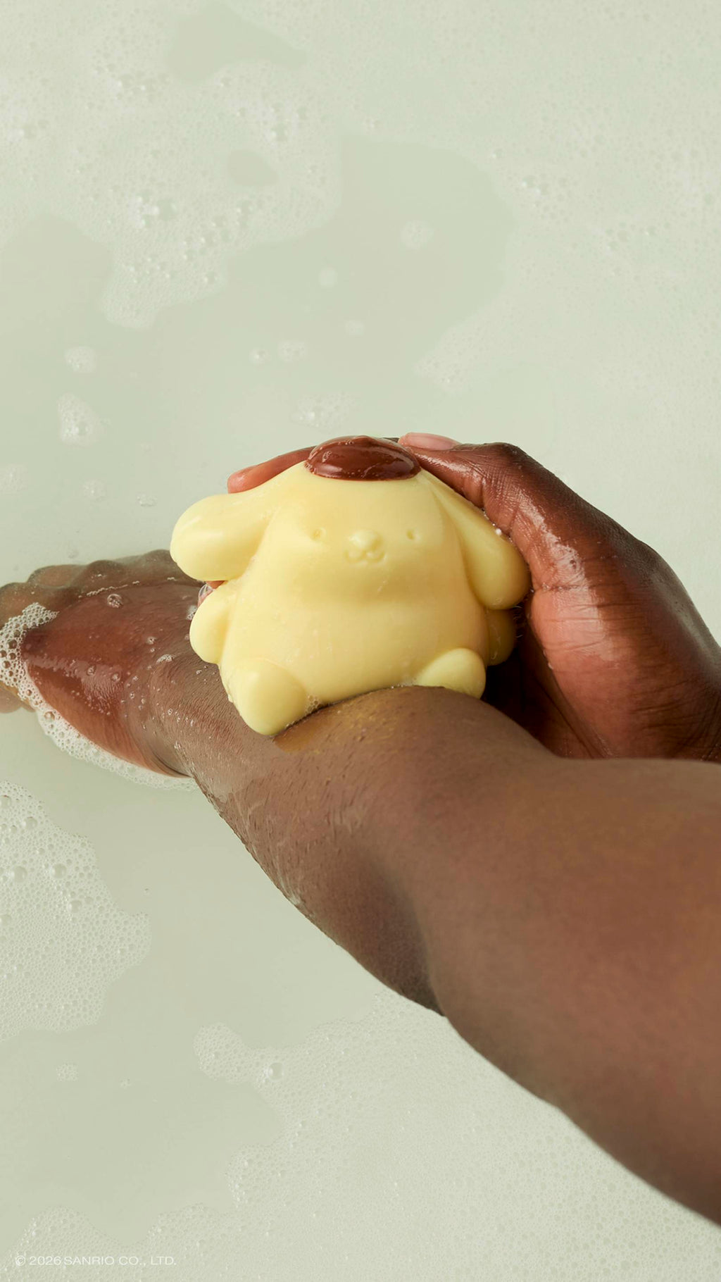 A model rubs a pale yellow Pompompurin shower jelly character against their arm. The glossy, dog-shaped character has a small brown beret and is held over a bath with soft bubbles on the surface.