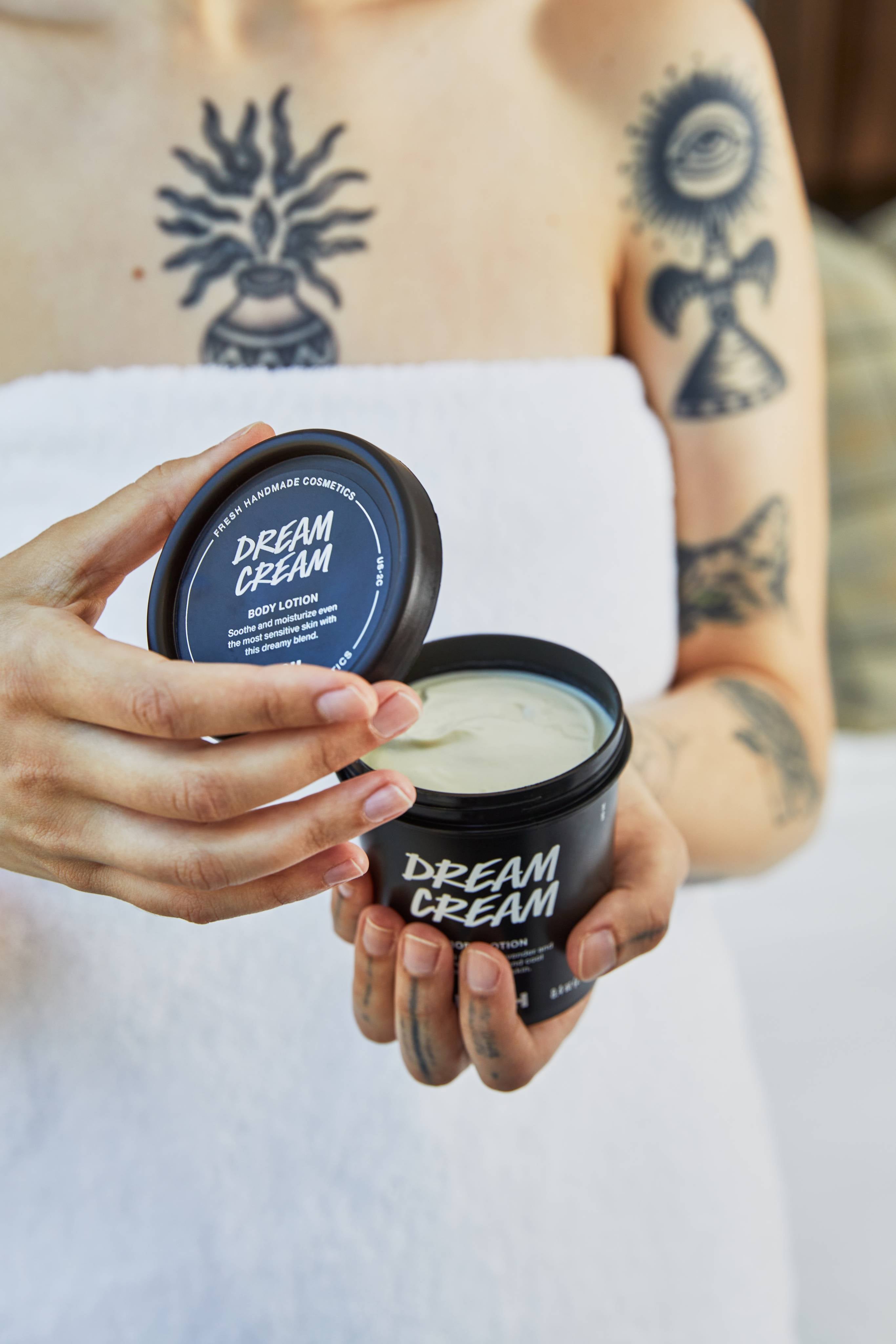 A tattooed person wrapped in a white bath towel holds an open black tub of Lush 'Dream Cream' body lotion, lifting the lid to reveal the pale, smooth cream inside.