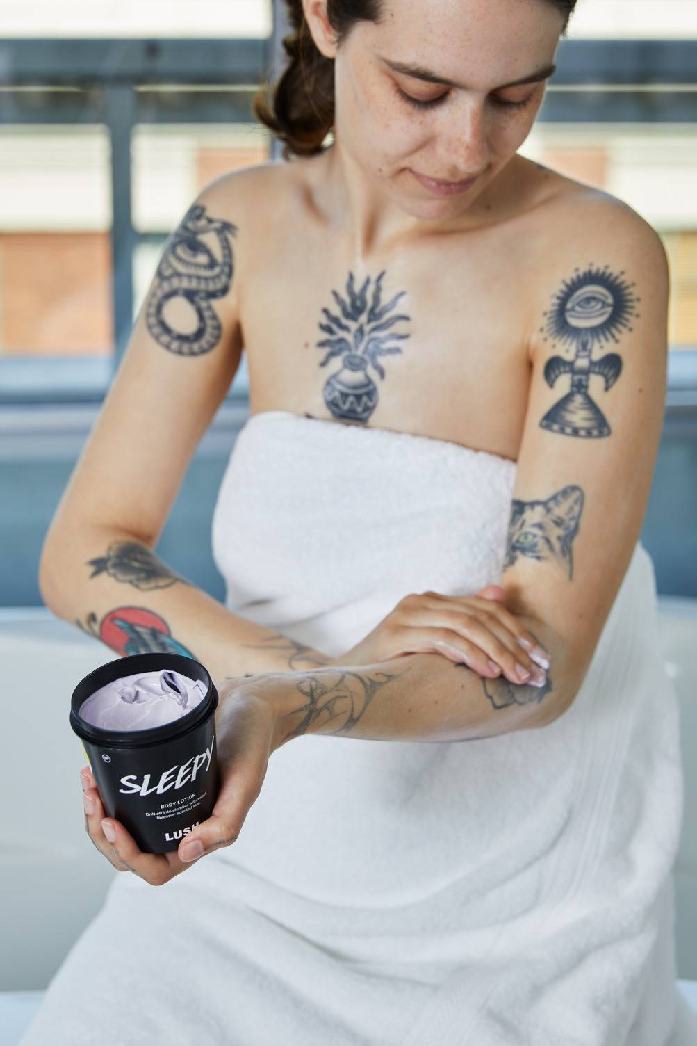 The image shows the model wrapped in a thick, white towel across their chest. The model has a few tattoos and is applying the Sleepy body lotion to their forearm.