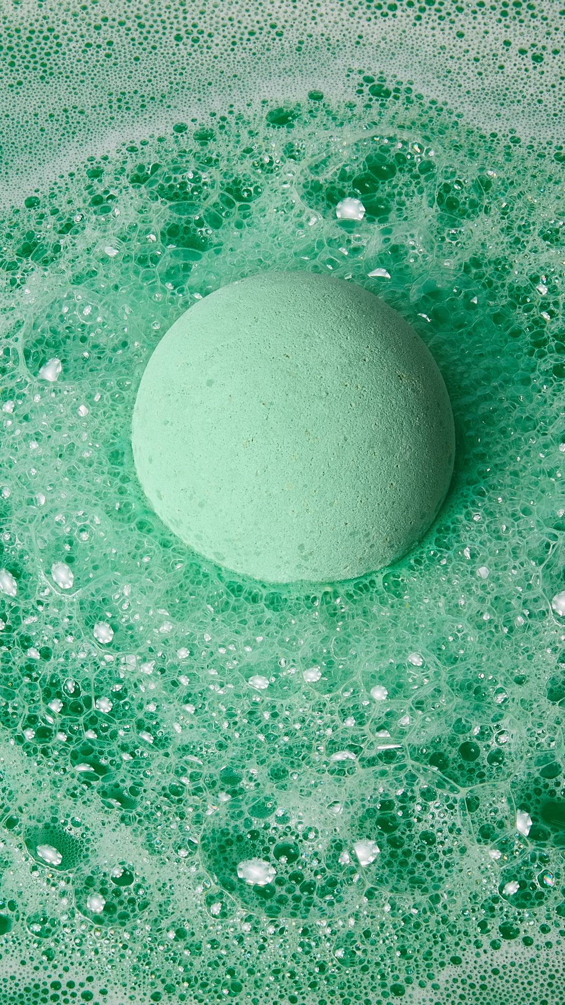 The image shows the pale green, round Avobath bath bomb floating on the surface of vibrant green bath water. Thick, foamy bubbles surround the bomb, creating a swirling texture across the water.
