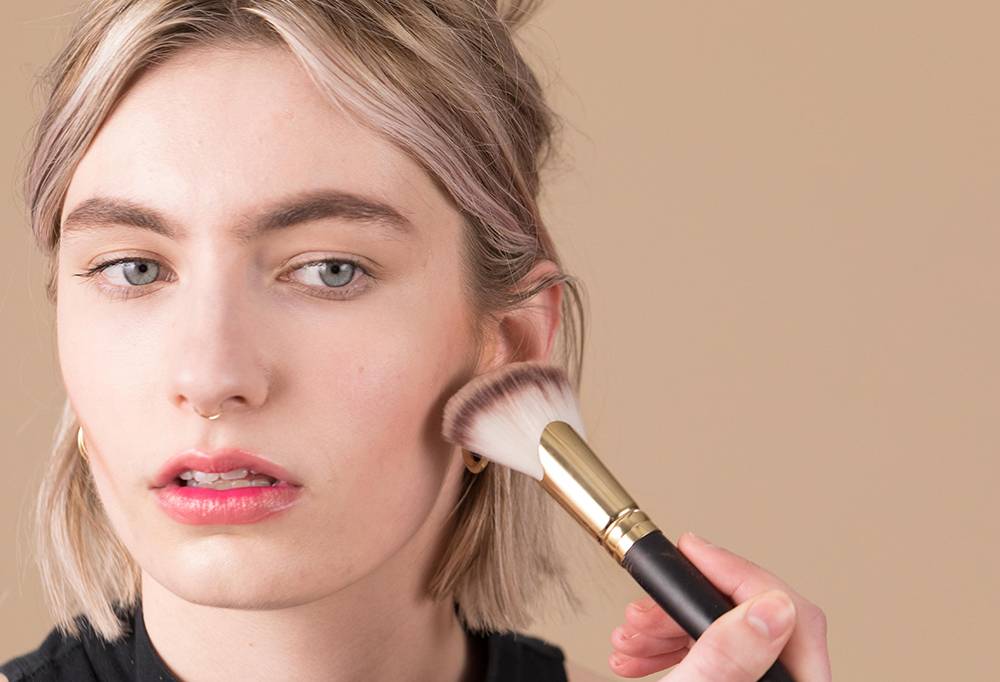 A person with blonde hair holds a Biggest Fan Brush close to their cheekbone, as if about to blend a contour in.