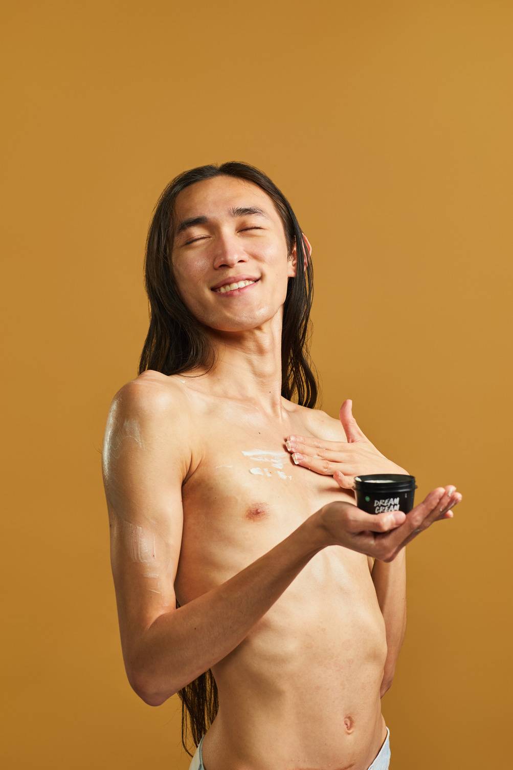 The model is topless and stands on a sandy, golden background. They have long hair and are closing their eyes and smiling as they apply the Dream Cream body lotion to their chest while holding the black pot in front of them.
