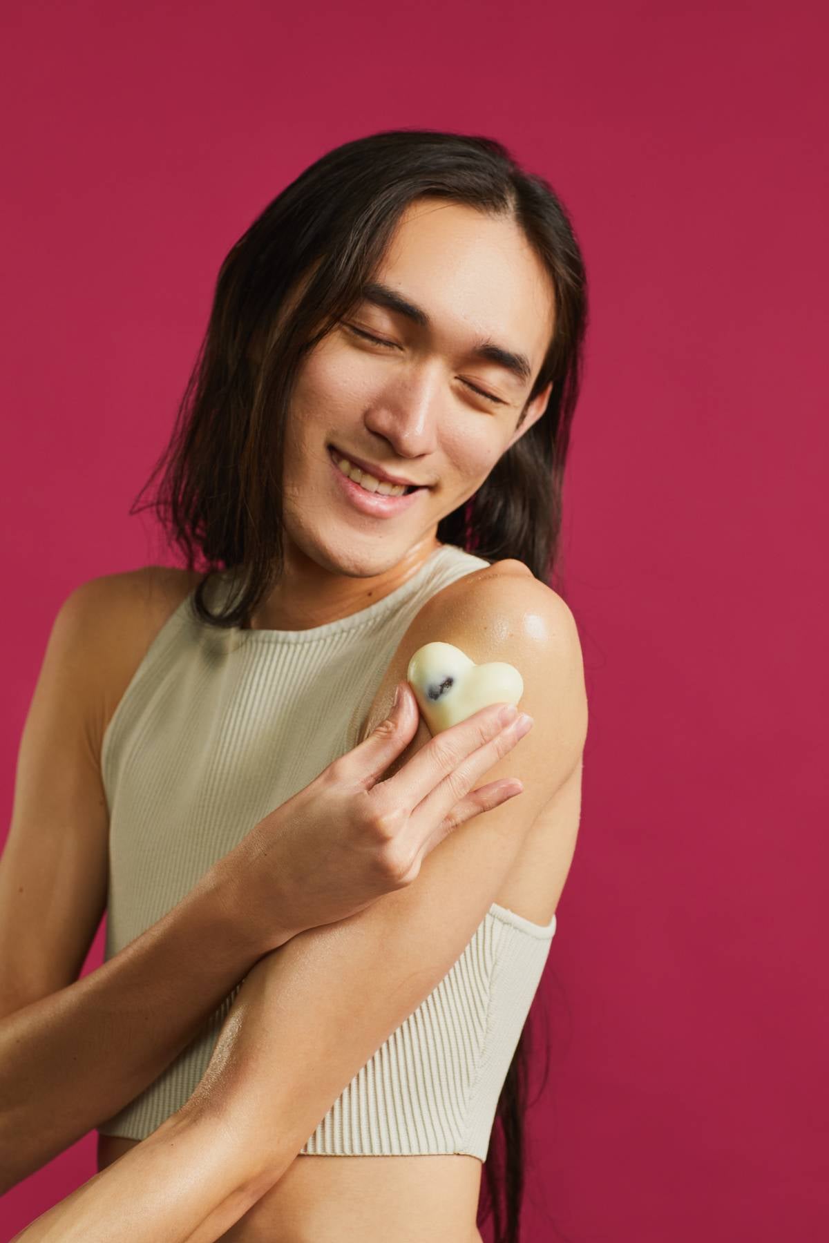The image shows the model on a rose-red background with a cream-coloured cropped top and long, dark hair. They use the heart-shaped Soft Coeur massage bar on their upper arm, moving it in gentle, circular motions.