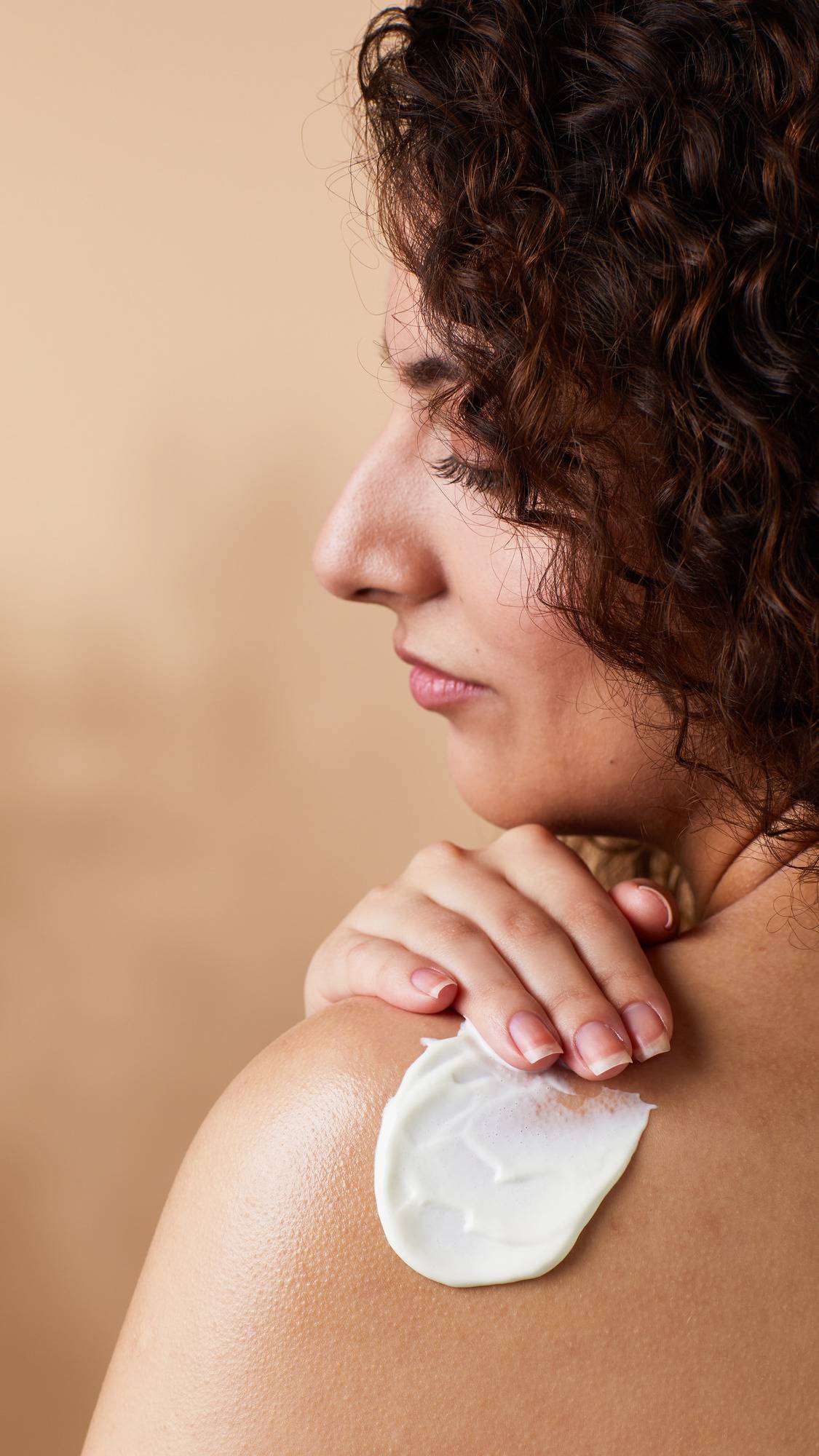 Close-up side profile of a person with dark curly hair smoothing a thick stroke of white cream onto their bare shoulder. Set against a warm, neutral beige background.