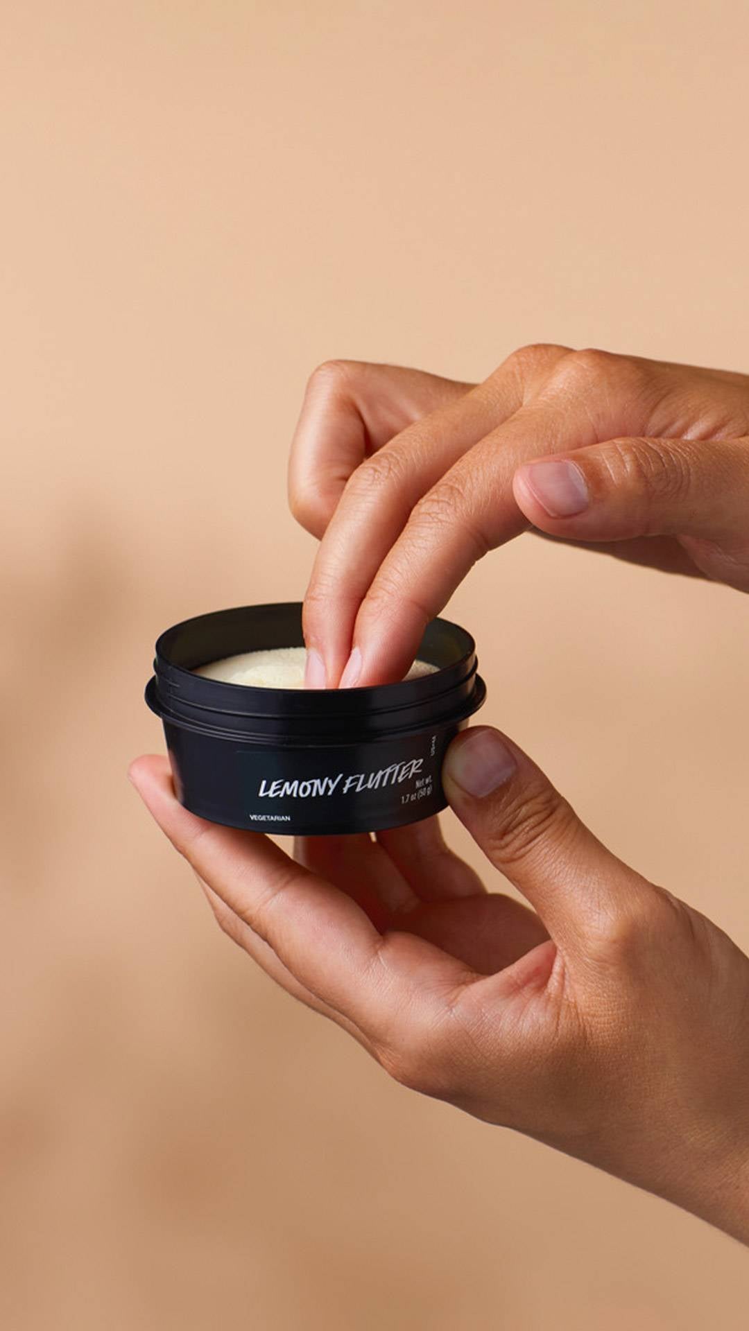 A pair of hands holds a small black tub of Lush "Lemony Flutter" cuticle butter against a tan background. Two fingers are dipped into the pale yellow product inside the pot, ready to apply.