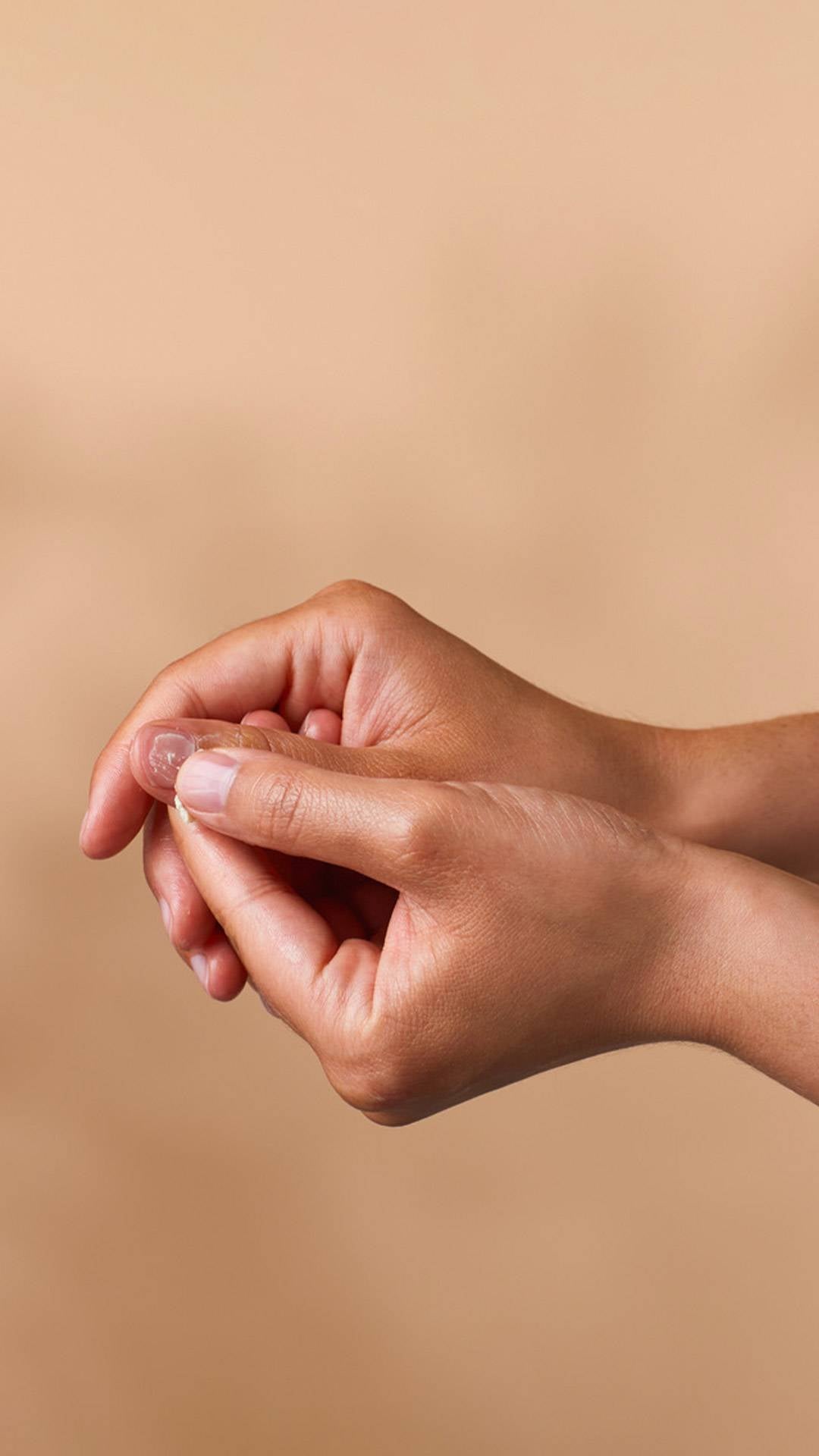 A close-up of hands against a tan background, demonstrating the final step of applying cuticle butter. The model uses their thumb to finish massaging the nourishing cream into the nail bed and surrounding skin for a hydrated, healthy look.