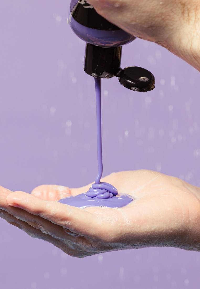 A thick stream of glossy, vibrant purple "Sleepy" shower gel pours from a black bottle, coiling smoothly into a wet palm against a soft lilac background with gentle water droplets.