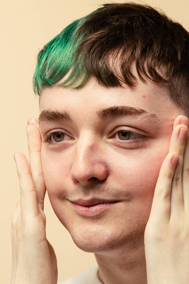 A close-up of a person with dark hair and a green dyed fringe gently touching their cheeks with both hands. Their skin looks smooth and moisturized, set against a warm, plain beige background.