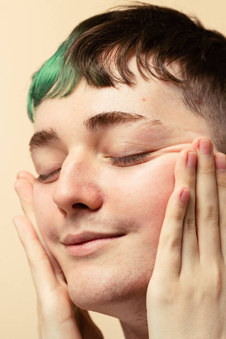 A close-up of a person with dark hair and a green dyed fringe gently touching their cheeks. Their skin looks smooth and moisturized, set against a warm, plain beige background.