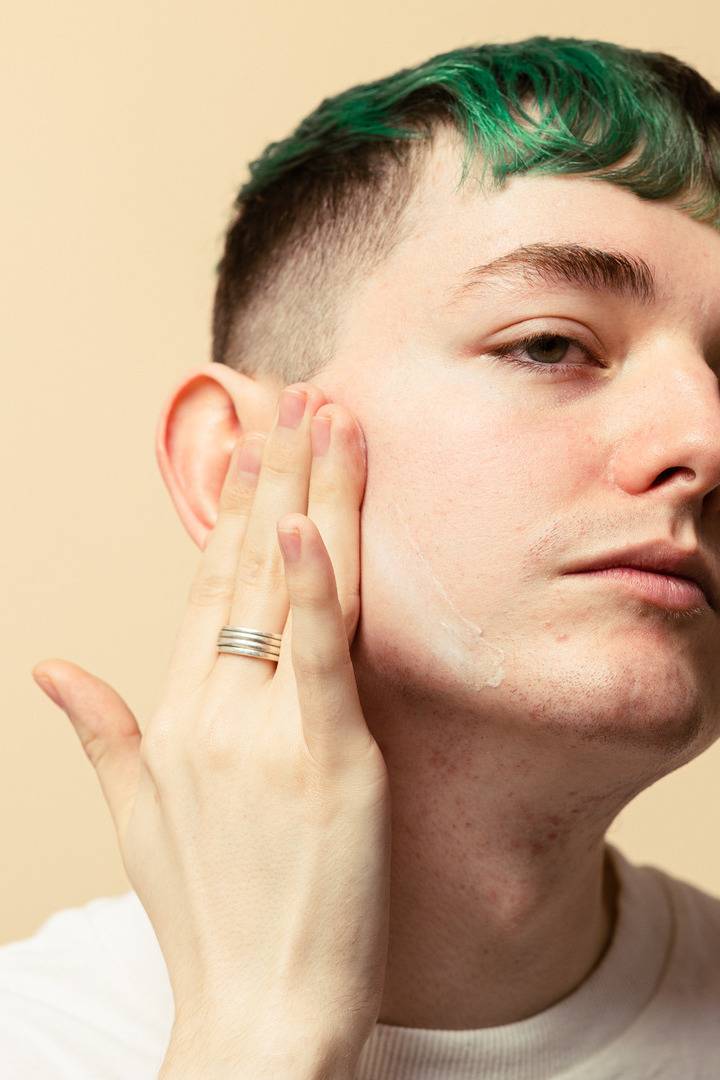 A close-up of a person with green dyed hair, eyes closed in relaxation, gently massaging the moisturizer into their temples and cheeks against a warm beige background.