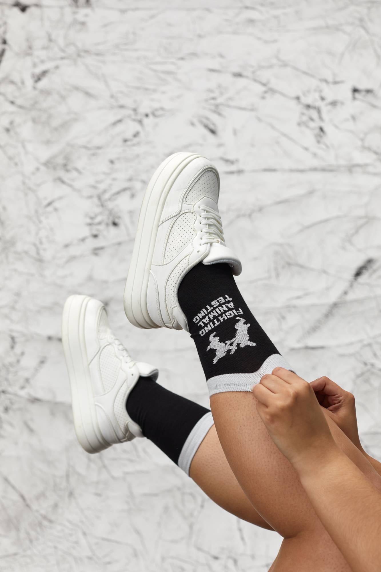 Model is pulling up the socks from the knee up as their legs are in the air. They wear white trainers on a marbled background.