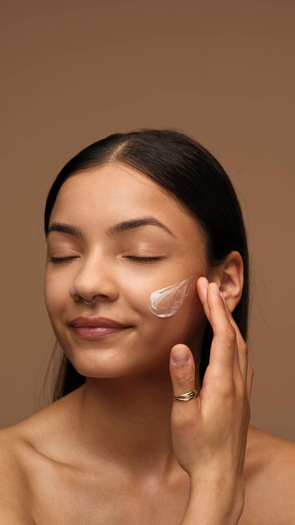A model with long dark hair and a septum piercing has their eyes closed while dabbing a patch of white Enzymion face cream onto their cheek. They are shown from the shoulders up against a solid brown background.