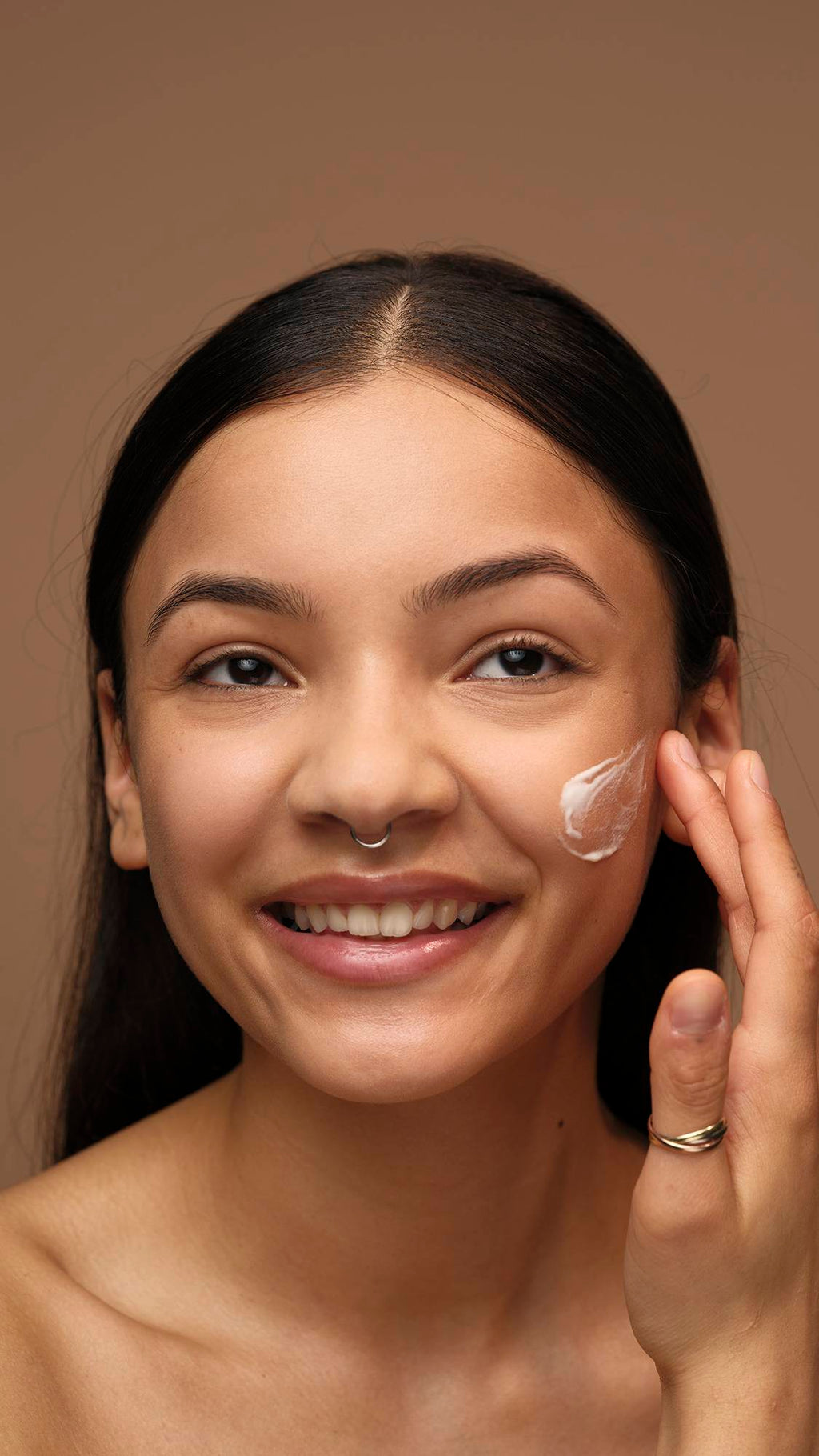 A model with long dark hair and a septum piercing smiles broadly while dabbing a patch of white Enzymion face cream onto their cheek. They are shown from the shoulders up against a solid brown background.