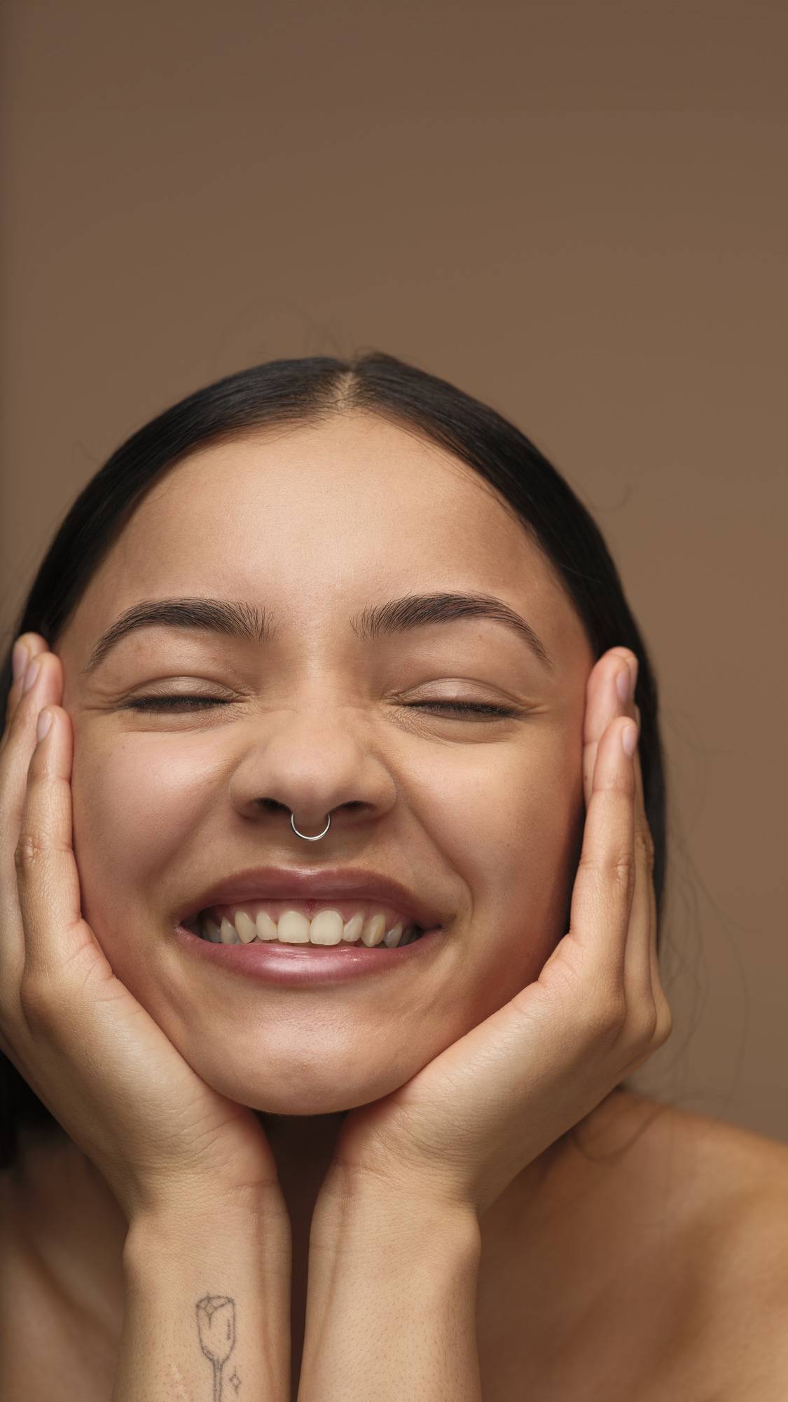 A model with long dark hair and a septum piercing smiles with their eyes closed, resting their freshly moisturised face in their hands. They have a small tattoo on their wrist and are posed against a solid brown background.