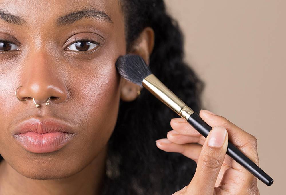 A close-up of person using Glow To Town, a blending/powder brush to blend highlight into their upper cheek area.