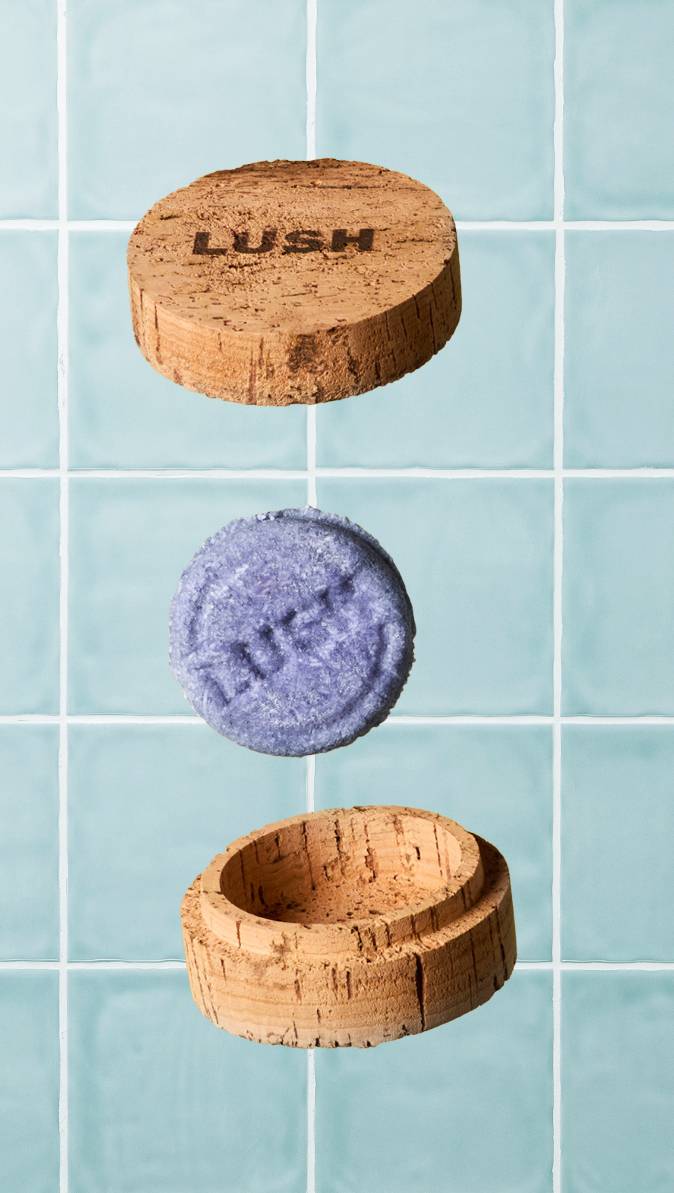 A textured purple Lush shampoo bar floats vertically between the two halves of an open, round cork storage pot. The top lid features a branded 'LUSH' stamp, set against a backdrop of light blue-green square wall tiles.