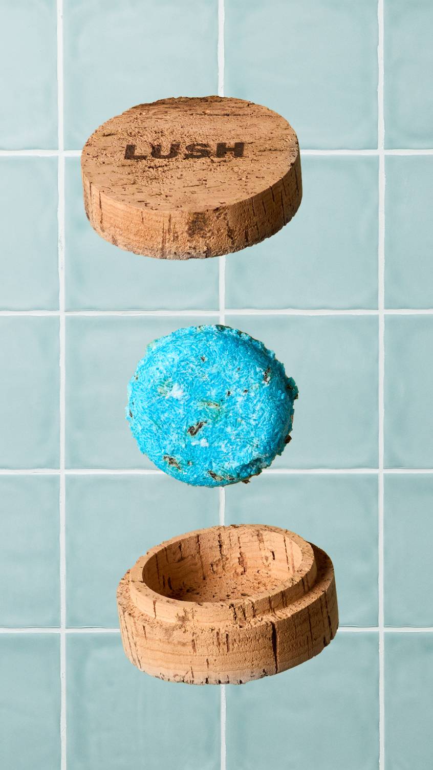 A bright blue circular shampoo bar floats in mid-air between the lid and base of an open, natural cork Lush container. The lid is branded with a dark 'LUSH' logo. Set against a background of pale blue square wall tiles.