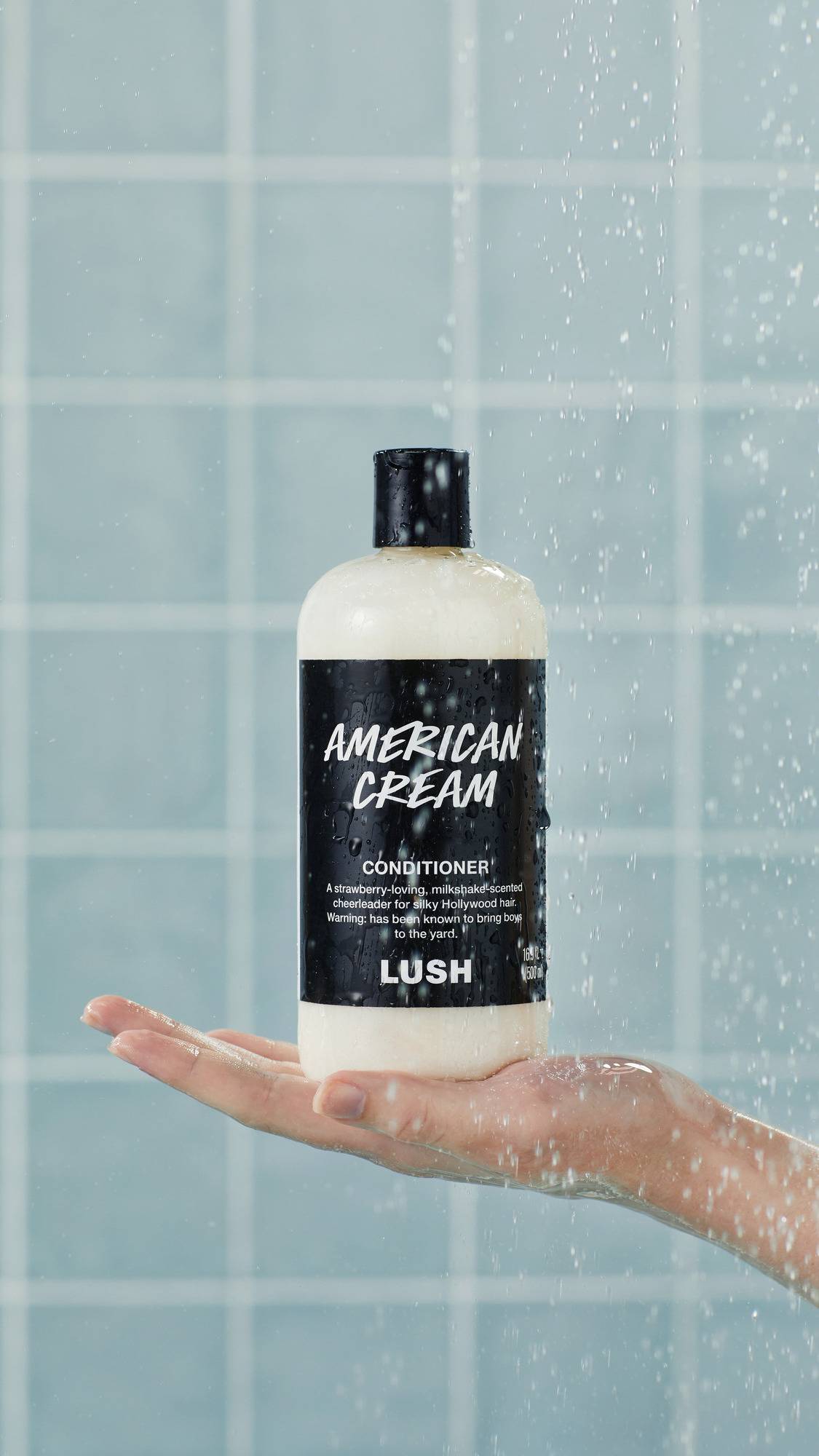 A bottle of Lush "American Cream" conditioner stands upright on an outstretched, wet hand in a shower. Water droplets spray across the black label and clear bottle against a background of glossy, light blue square tiles.