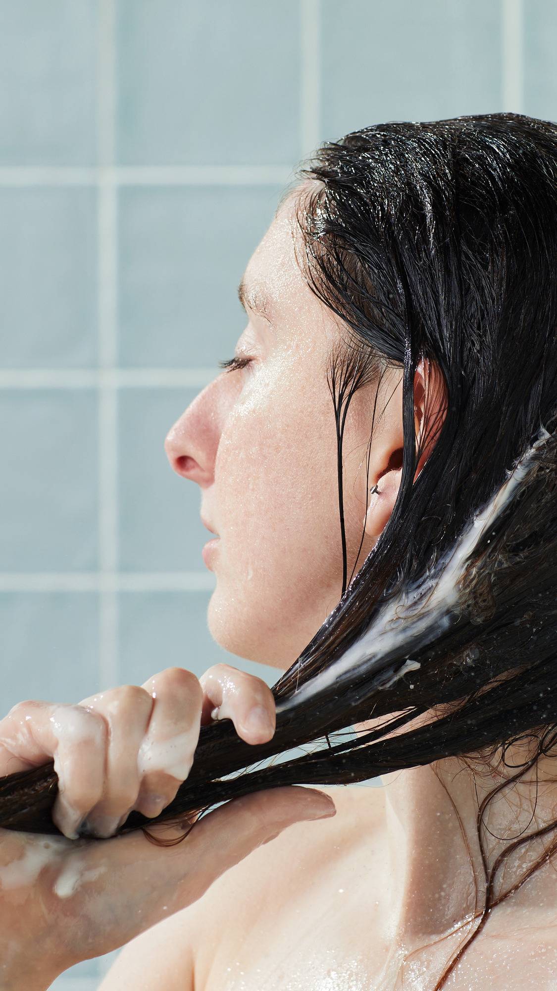 A side-profile view of a model with eyes closed, running the white Lush "American Cream" conditioner through their long, dark hair in the shower. Their hands are coated in product against a backdrop of glossy, light blue square tiles.