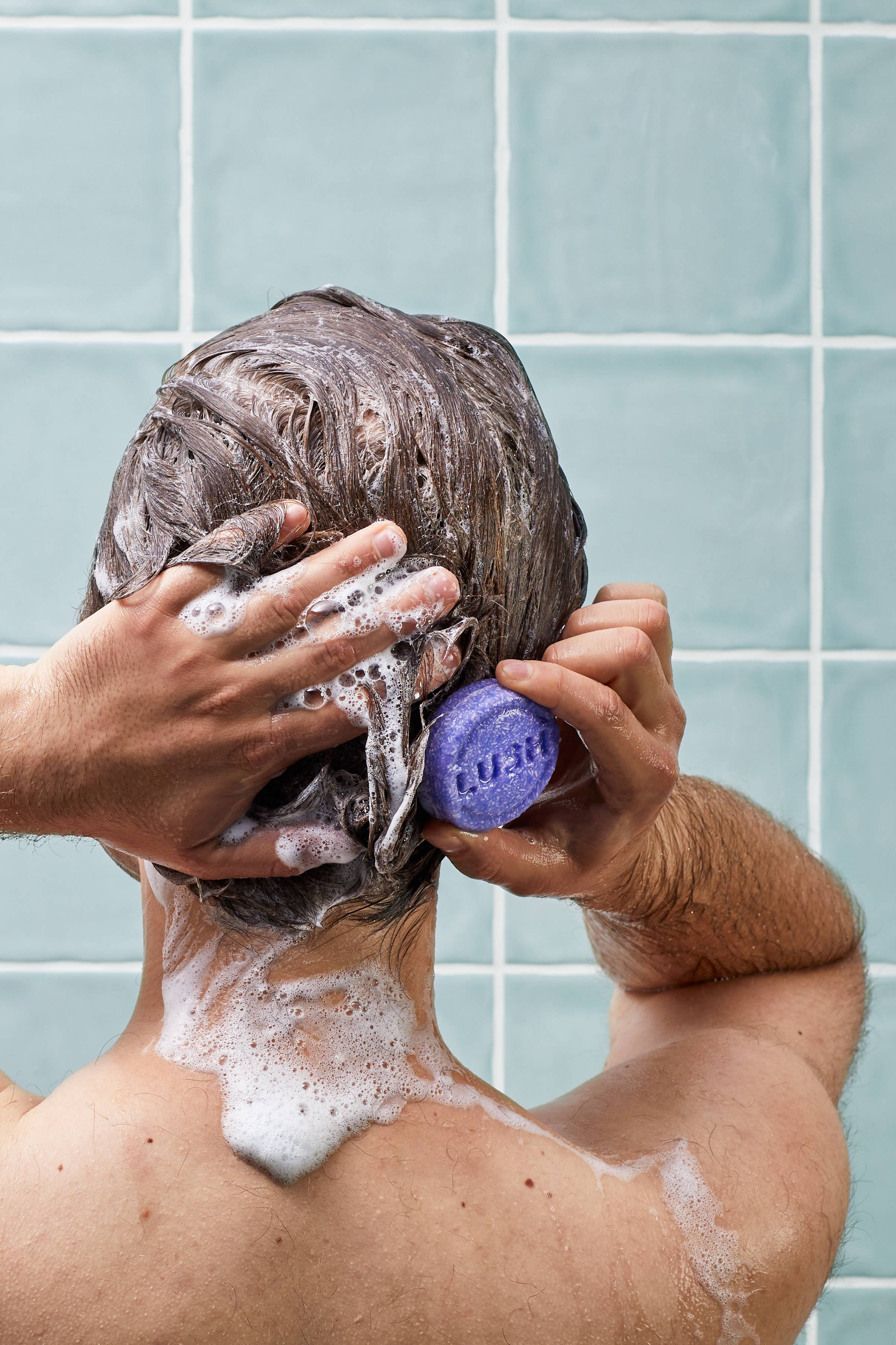A man in the shower washes his hair using a circular purple Lush solid shampoo bar, creating a white lather. He is shown from behind against a background of light blue-green square wall tiles.
