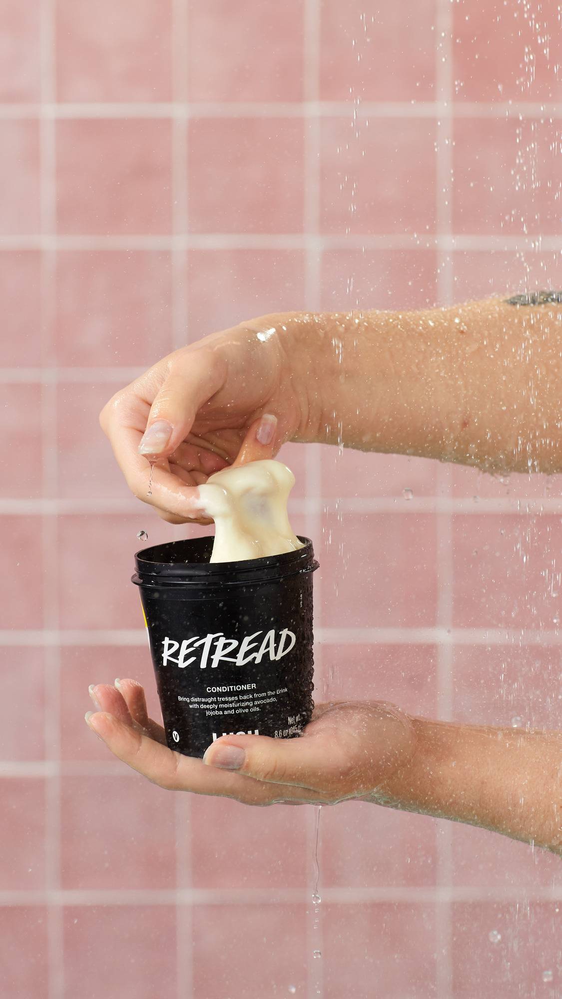 The image shows a model's hand scooping a thick dollop of cream-coloured conditioner out of a black Lush "Retread" pot. They are holding the container in a shower with falling water droplets against a background of pink square tiles.