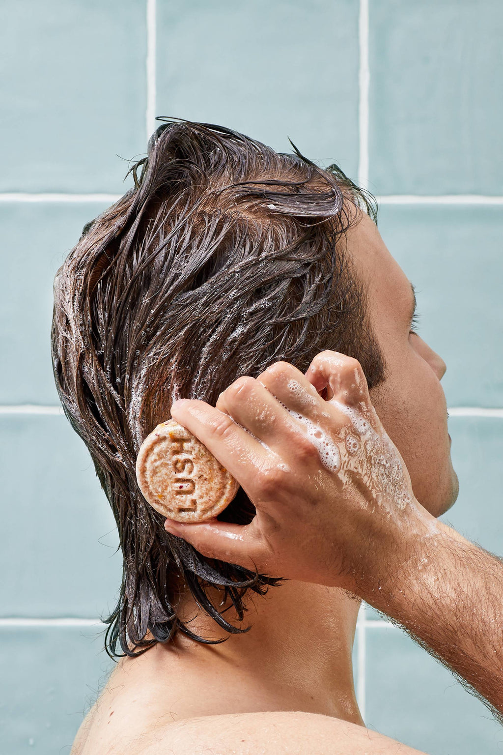A person with dark, wet hair glides a circular, textured cream Lush shampoo bar directly over their head, creating a light foam. The scene is set in a shower against a background of pale blue square wall tiles.
