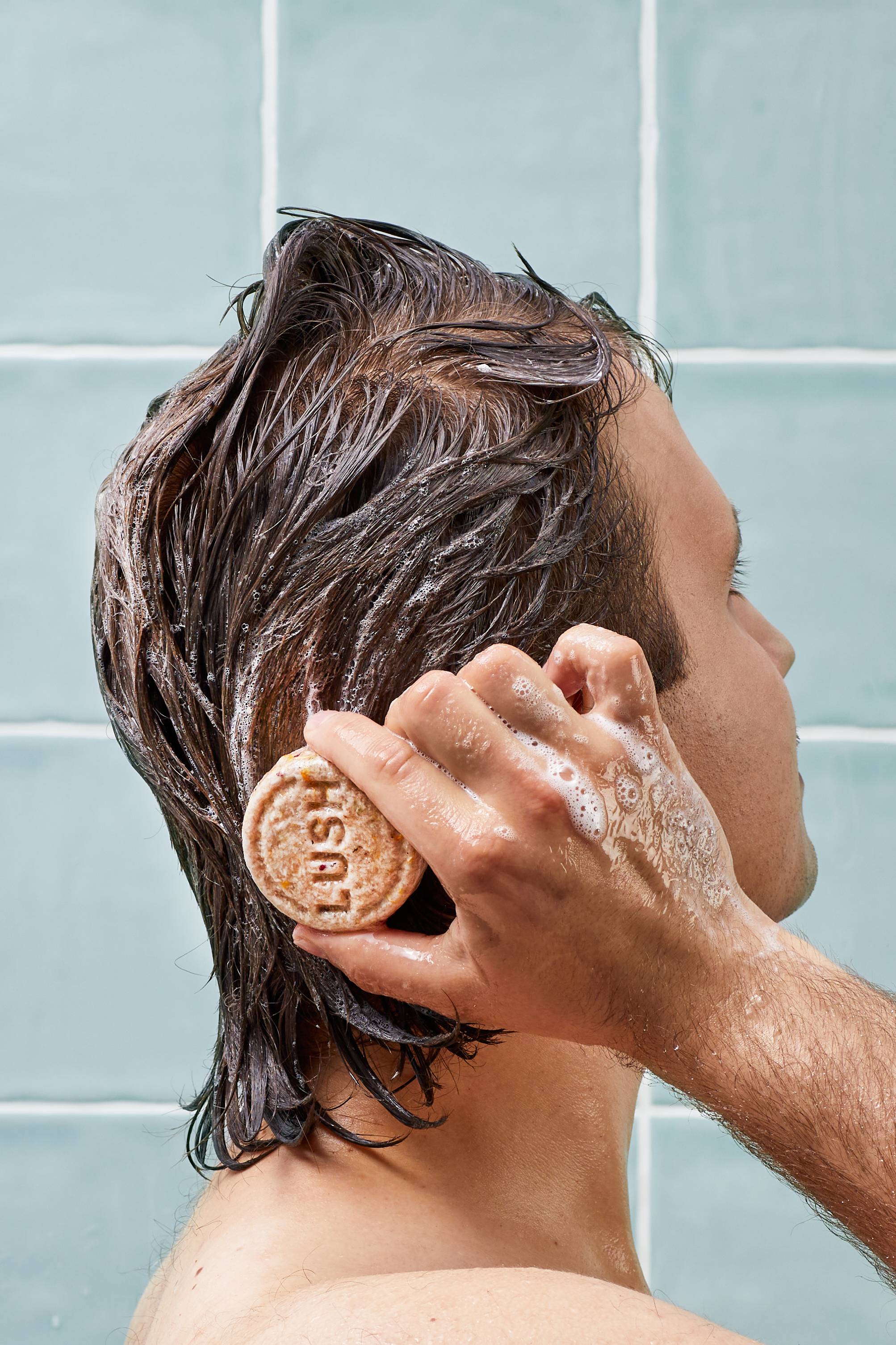 A person with dark, wet hair glides a circular, textured cream Lush shampoo bar directly over their head, creating a light foam. The scene is set in a shower against a background of pale blue square wall tiles.