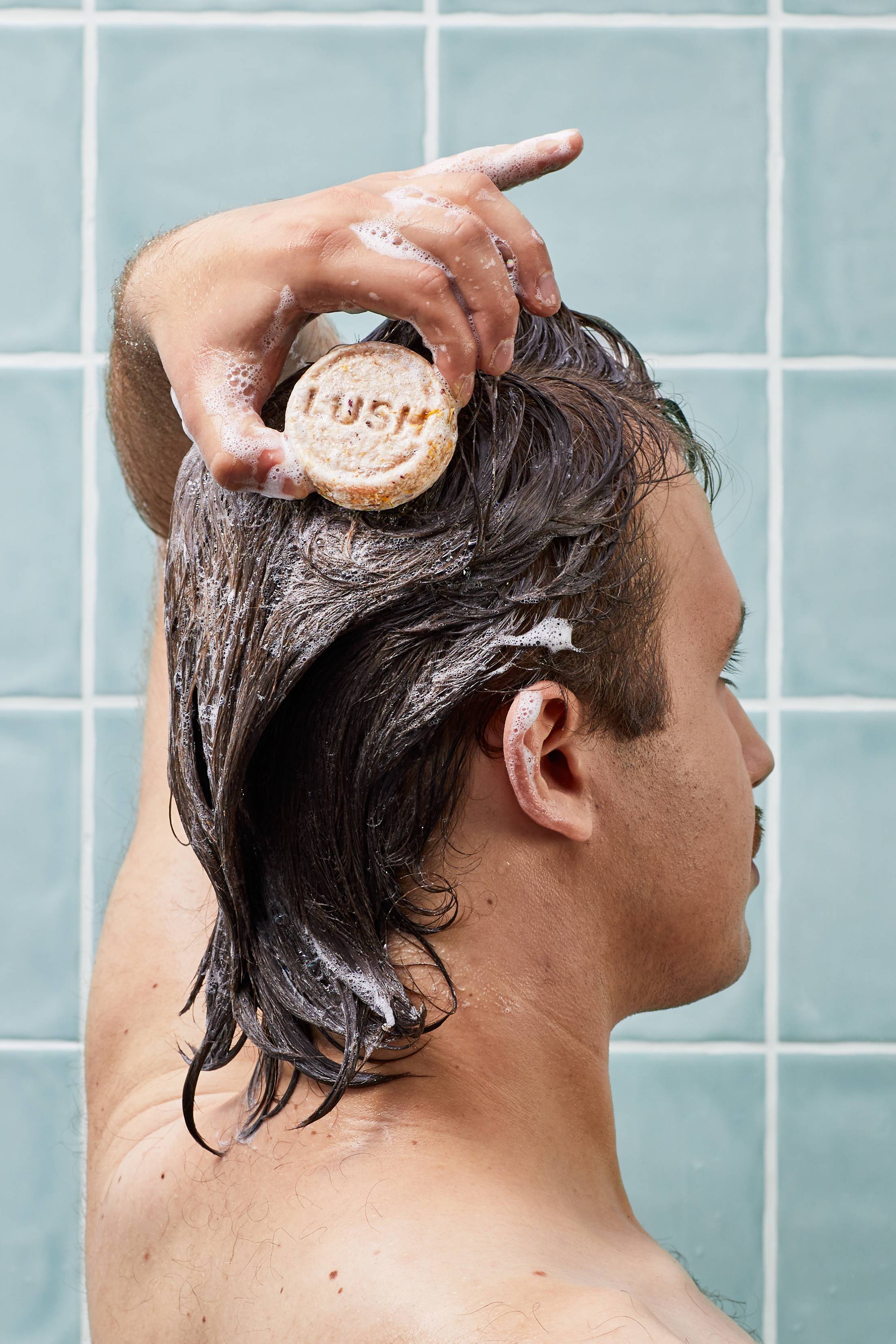 A person with dark, wet hair glides a circular, textured cream Lush shampoo bar directly over their head, creating a light foam. The scene is set in a shower against a background of pale blue square wall tiles.