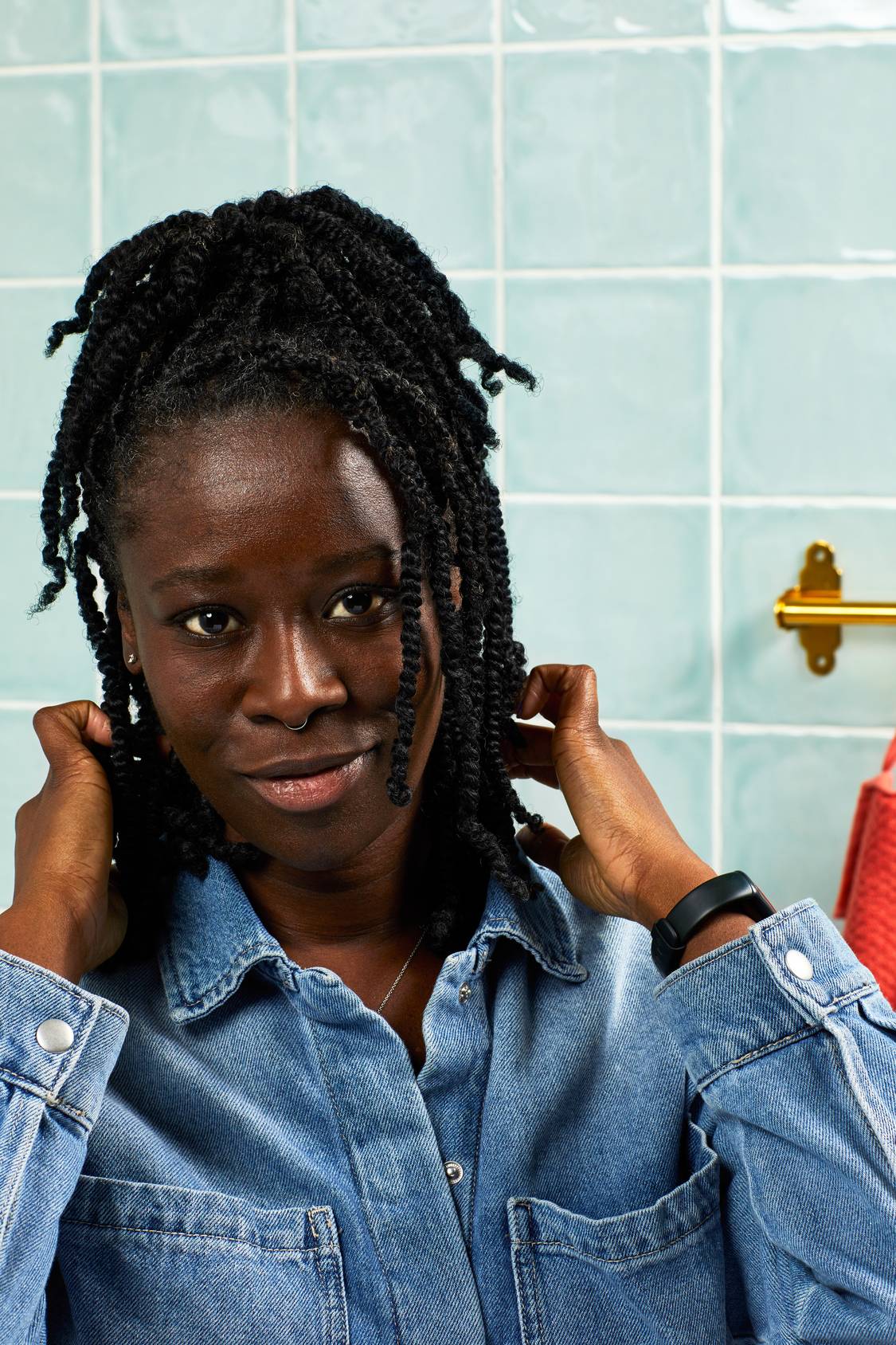 A smiling person with braided hair poses against a backdrop of light blue bathroom tiles. Wearing a denim shirt, they hold the ends of their hair with both hands, looking directly at the camera.